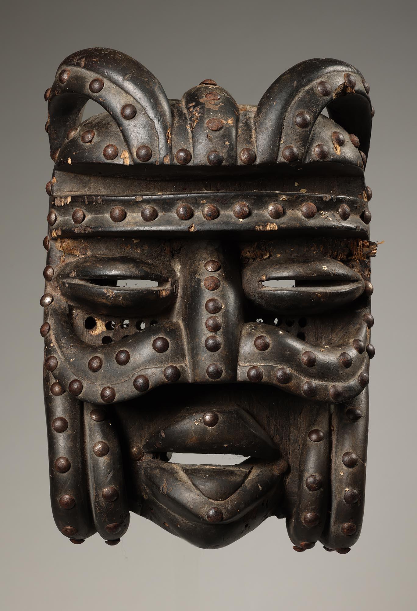 Masque en bois sombre des peuples bété, avec moustache recourbée, défenses recourbées par la bouche et cornes recourbées sur le dessus.  Bouche ouverte avec langue et punaises métalliques, Côte d'Ivoire, Afrique. Bonne patine foncée et clous oxydés