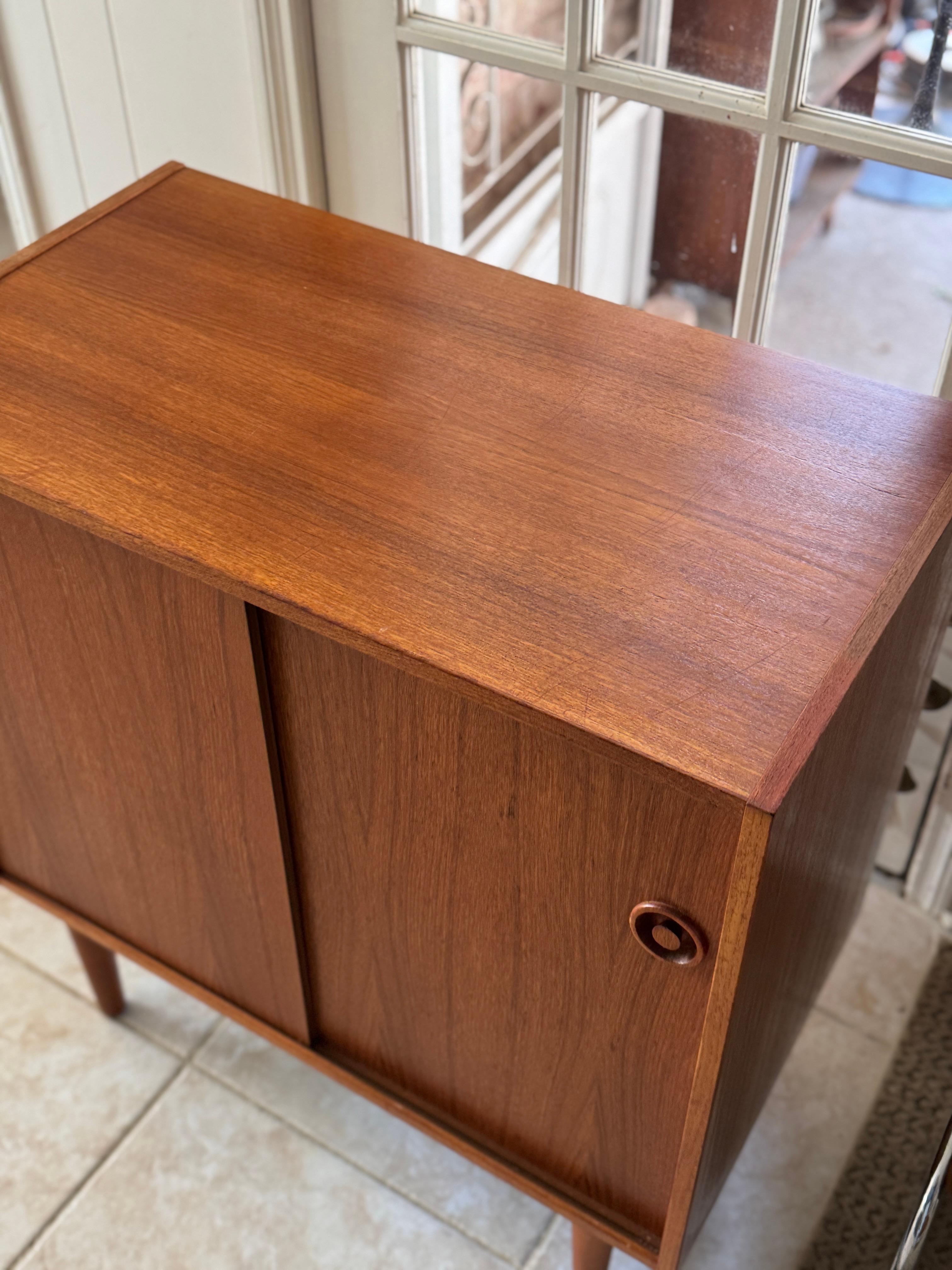 Classic Danish mid century modern teak record cabinet with sliding doors, circa 1960s. The sliding doors reveal ample space perfect for storing vinyl records, books, or barware. Finished with recessed circular pulls and warm teak grain, it’s a