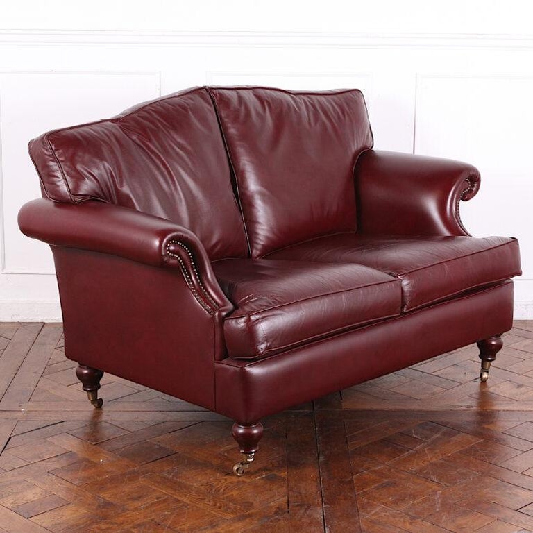 Classic rolled-arm English-made two seat sofa in oxblood leather. Brass nail head details; raised on turned mahogany bun feet with brass castors. Leather in excellent condition. Purchased from a large estate in England. The suite was situated in the