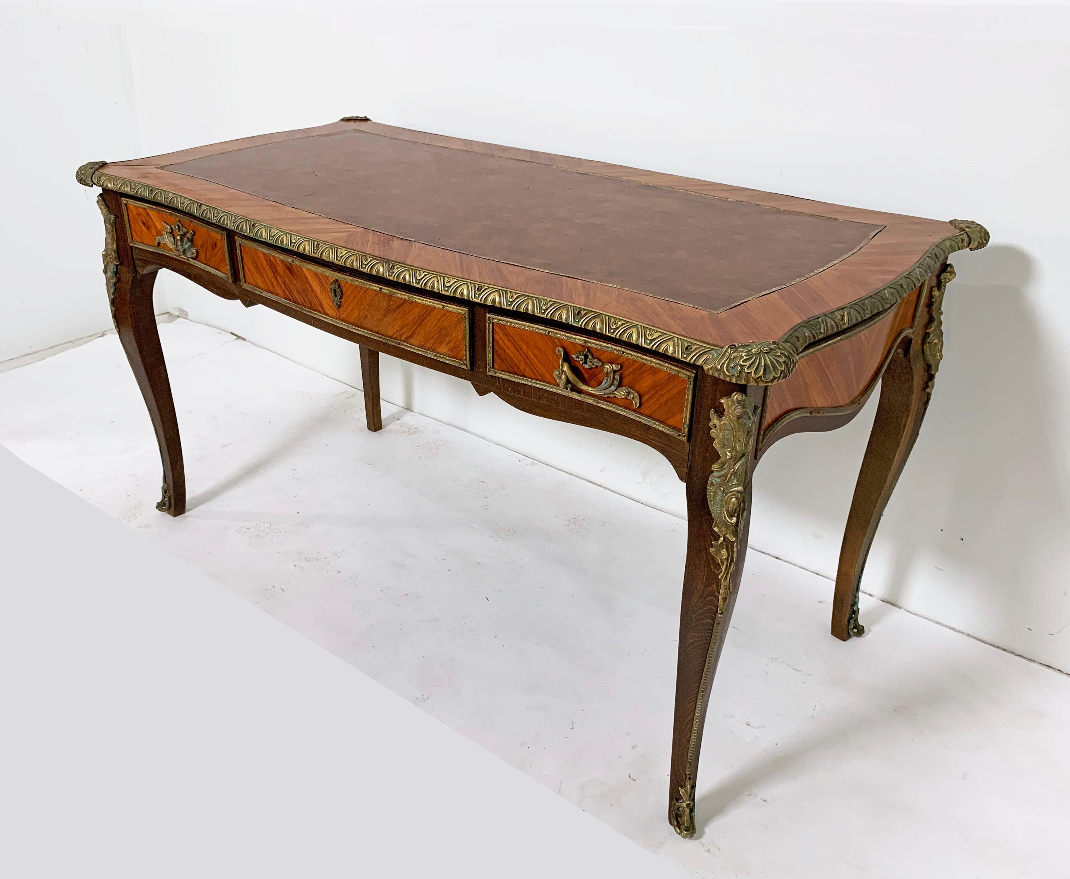 A Classic French Louis XV “bureau plat” writing desk in ribbon mahogany and walnut with brass ormolu and leather writing surface panel, circa 1950s.