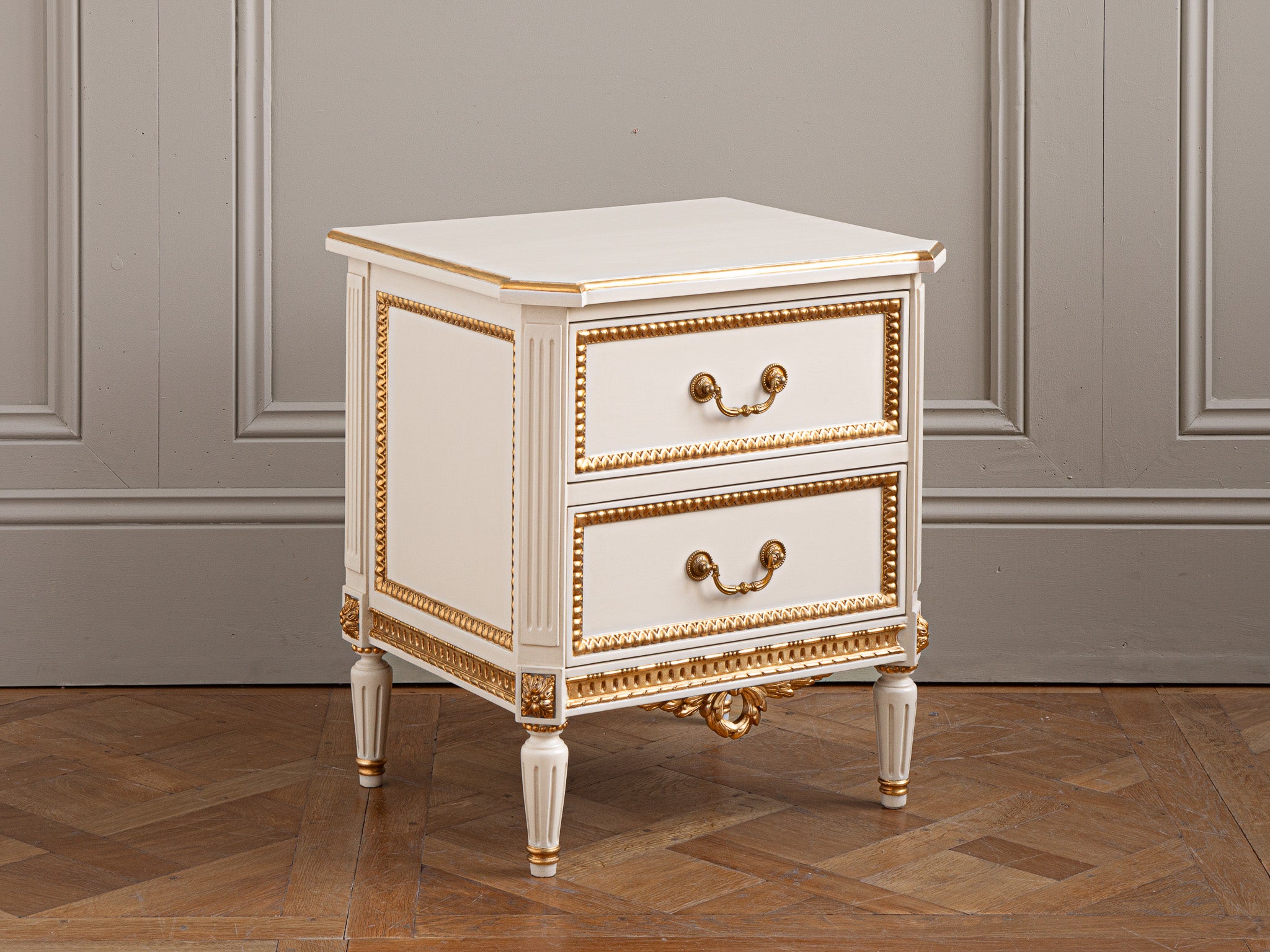 A Pair of bedside tables made by artisan furniture specialists - La Maison London. 
The Bedsides have been hand made in wood, in the classic Louis XVI style, and finished in an ecru white patina with gilt work to highlight the accents of this