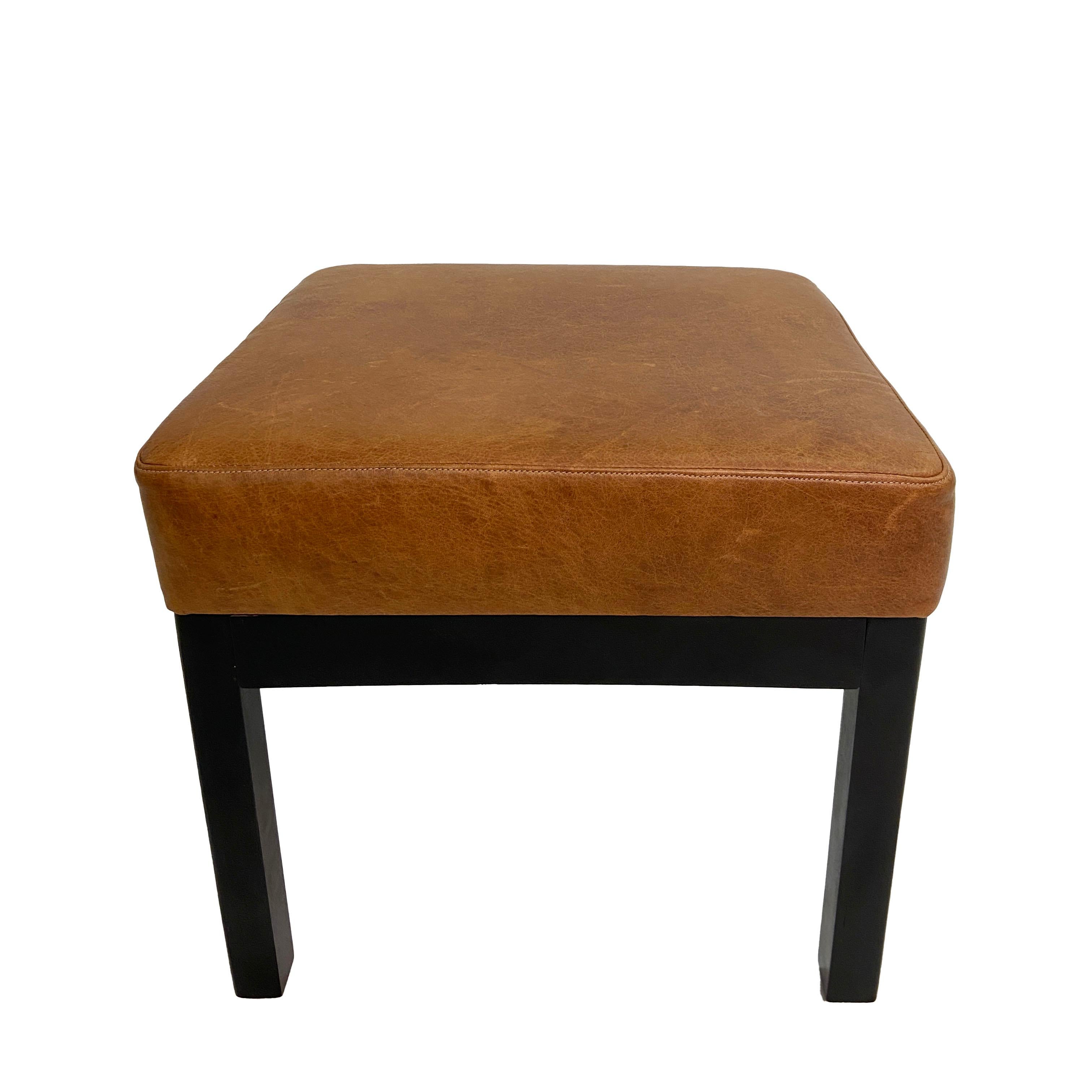 A pair of classic square low stools with cushions upholstered in handsome and mildly distressed brown leather. The solid wooden frames have been refinished in ebony. These modifications have been newly done and in excellent condition. Tuck them