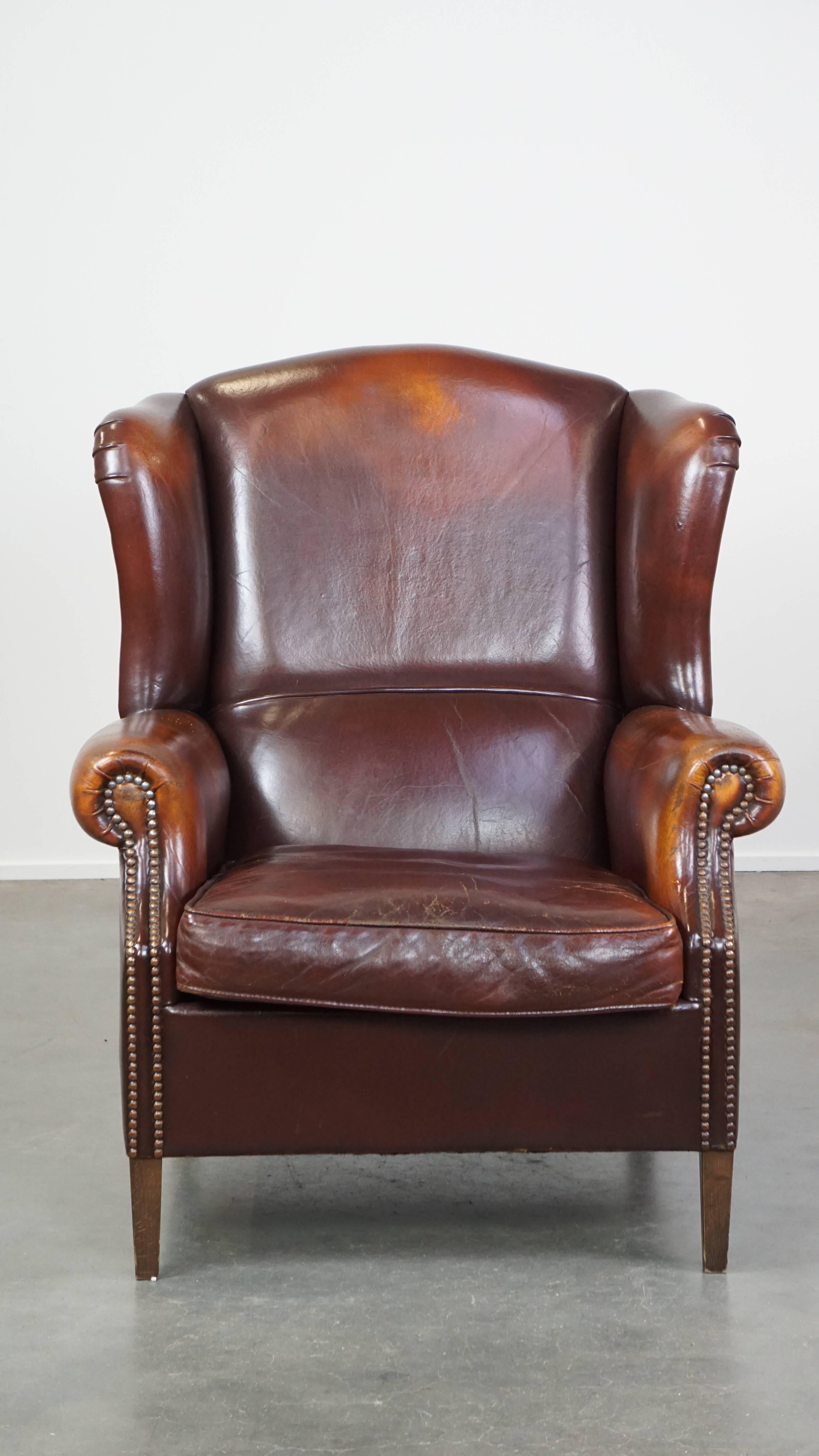 Ce fauteuil à oreilles en cuir de mouton incarne le style anglais classique grâce à sa combinaison de luxe et de confort intemporels. Il a une couleur riche et profonde et est terminé par des clous décoratifs. Le fauteuil est doté d'un coussin