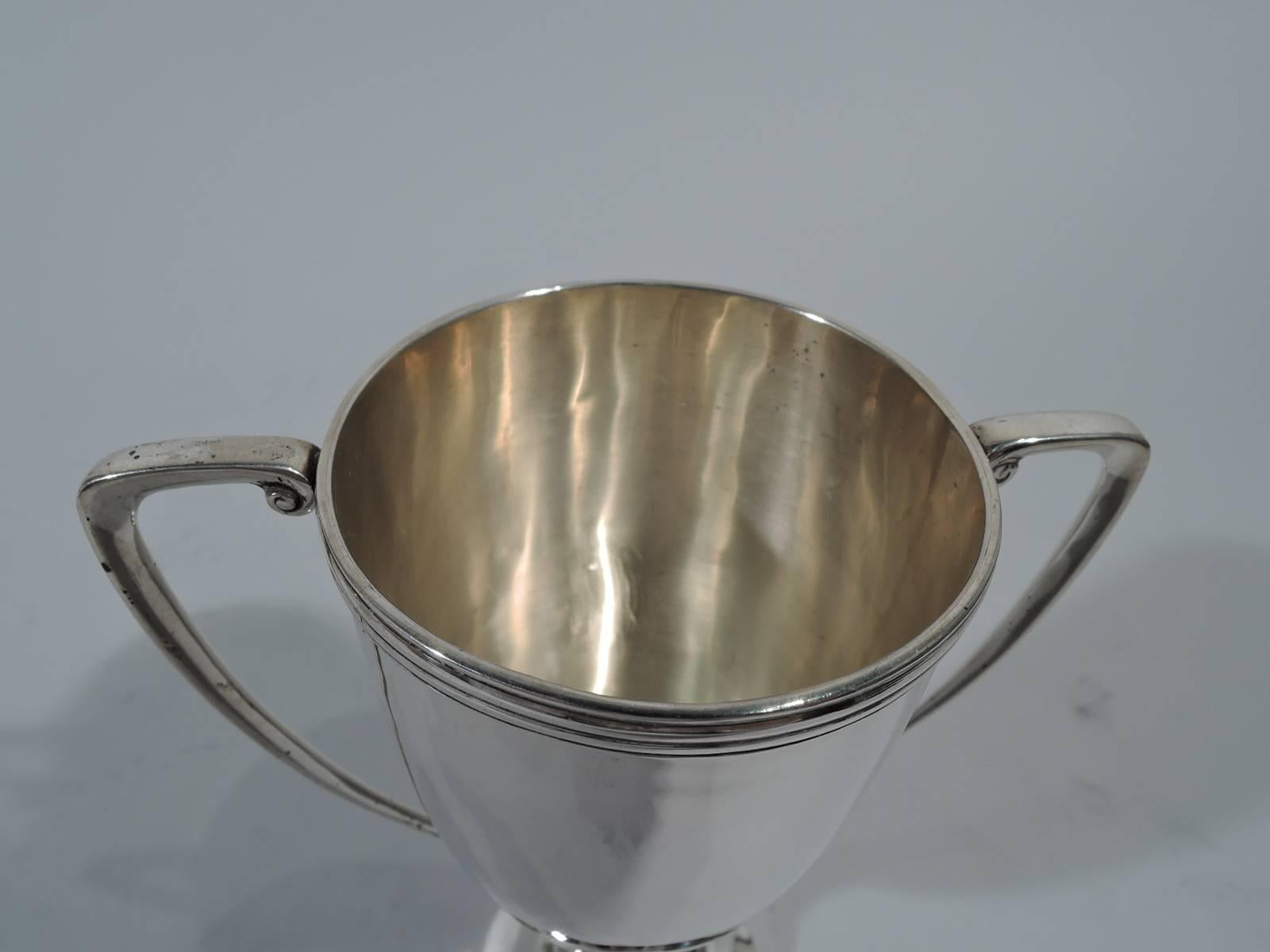Classic silver trophy cup. Made by Hamilton & Co. in Calcutta. Tall and narrow bowl with reeded rim. Domed foot. Scrolled-bracket handles with volute scrolls. Traditional form gracefully rendered in small-scale. Hallmarked. Weight: 4.5 troy ounces.