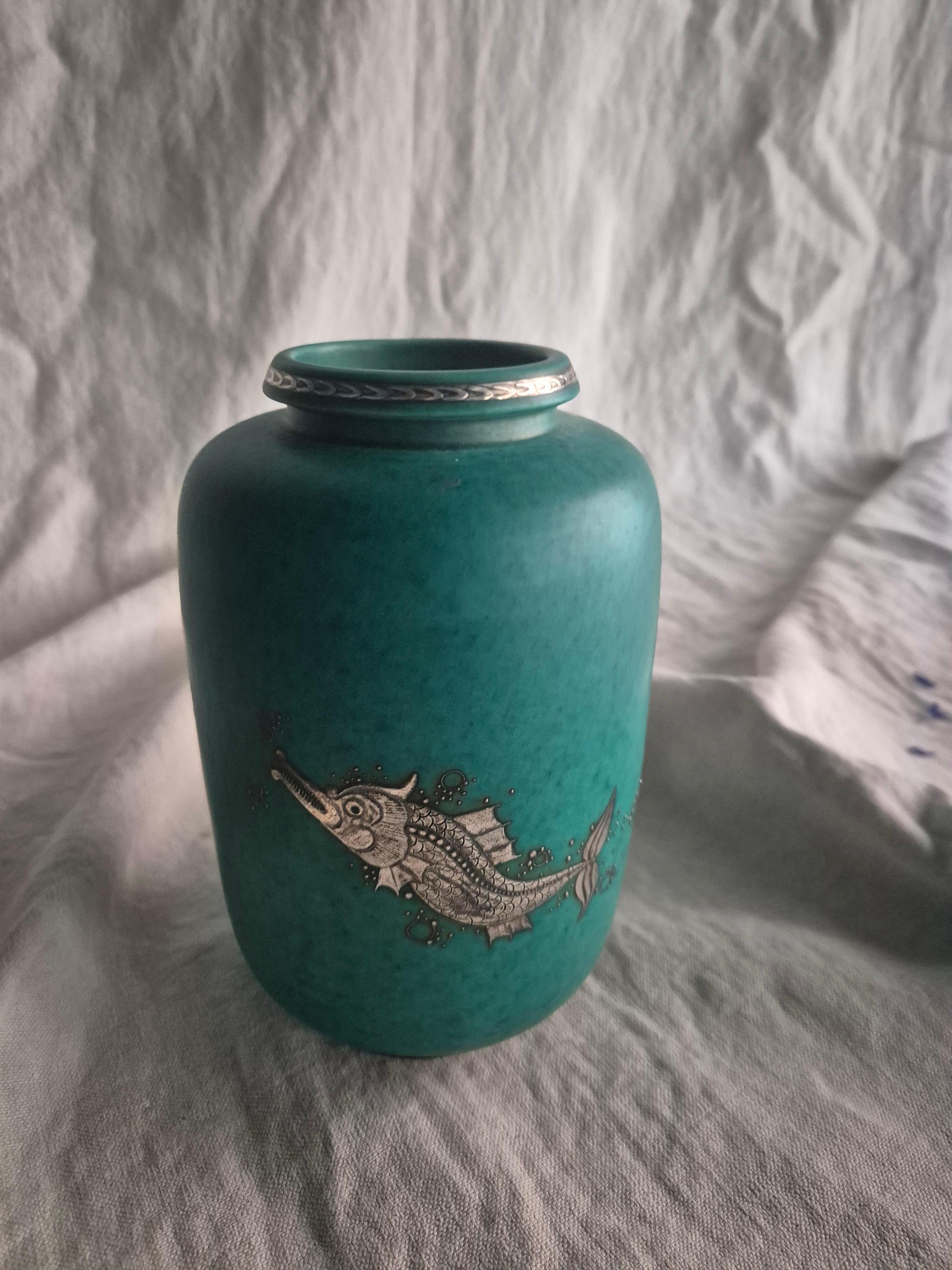 A Swedish modern classic, stoneware vase in signature green glaze with decor of a mythical fish at play, and stripe at the rim in silver. Designed by Wilhelm Kåge from the ‘Argenta’ series for Gustavsberg, Sweden, 1938, (year letter H). 


Signed in