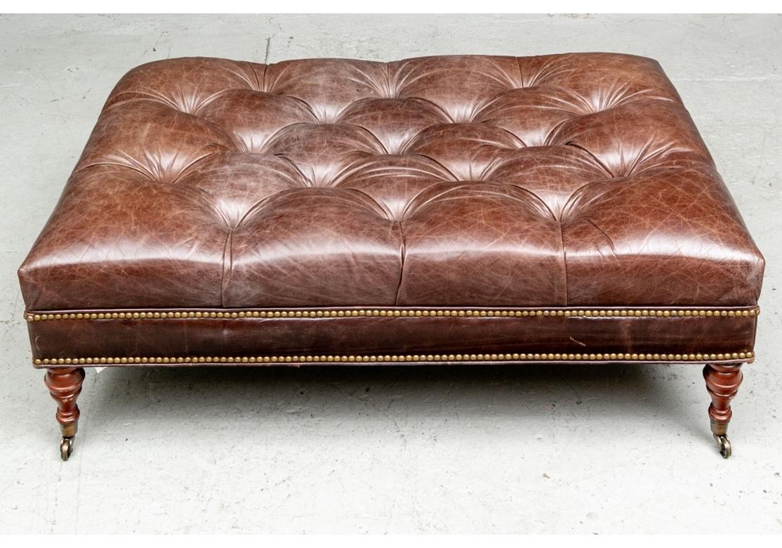 A large scale good looking and useful size brown tufted leather ottoman with brass nail head trim on the skirt. Raised on brown stained turned legs on brass casters. 
Measures: length 48