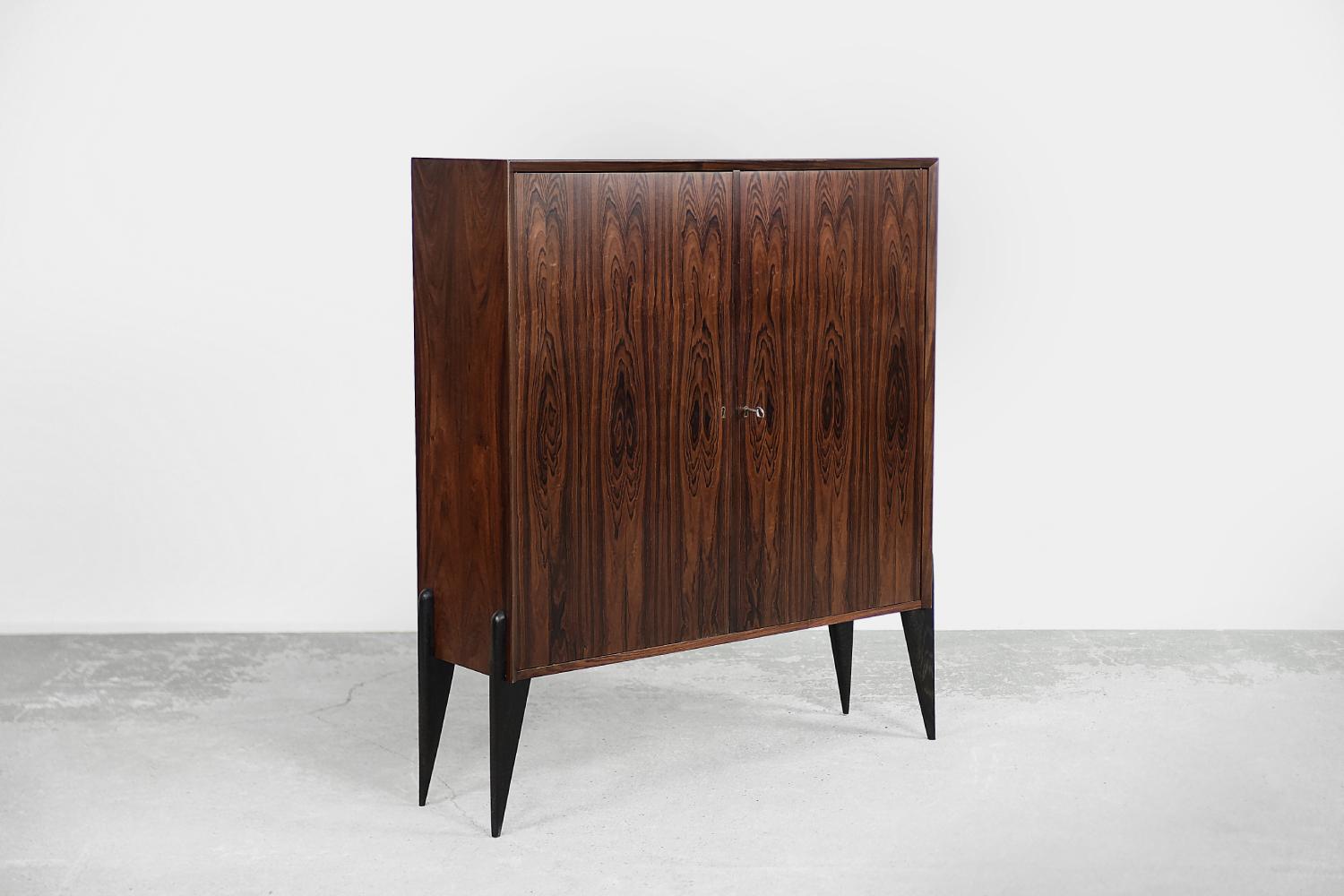 This modernist, narrow cabinet was made in Denmark during the 1960s. It was finished with precious rosewood with strongly marked grains. It has a pair of lockable doors. Inside, on both sides there are shelves whose height can be adjusted. The base