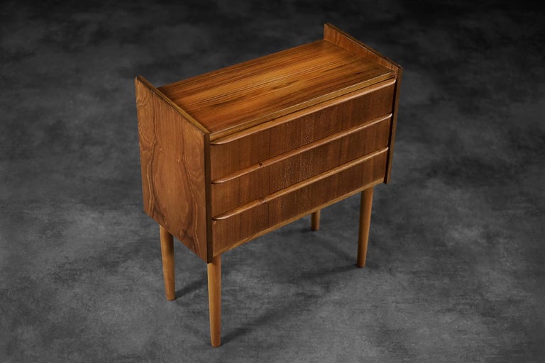 This small modernist cabinet was made in Denmark during the 1960s. It is made of teak wood in a warm shade of brown. It has three drawers with wooden handles. The base is turned legs made of solid wood. It was made in accordance with the tradition