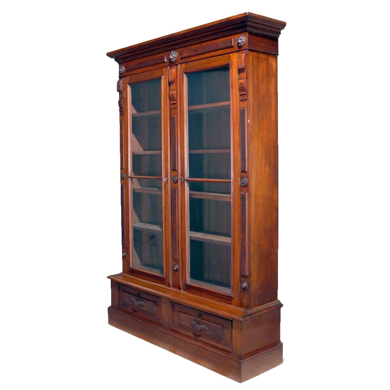 Classical walnut bookcase features case with burl and carved lion head reserves and two glass-front doors opening to reveal adjustable shelf interior and over two lower drawers, locking and with key, circa 1890.

Measures: 85