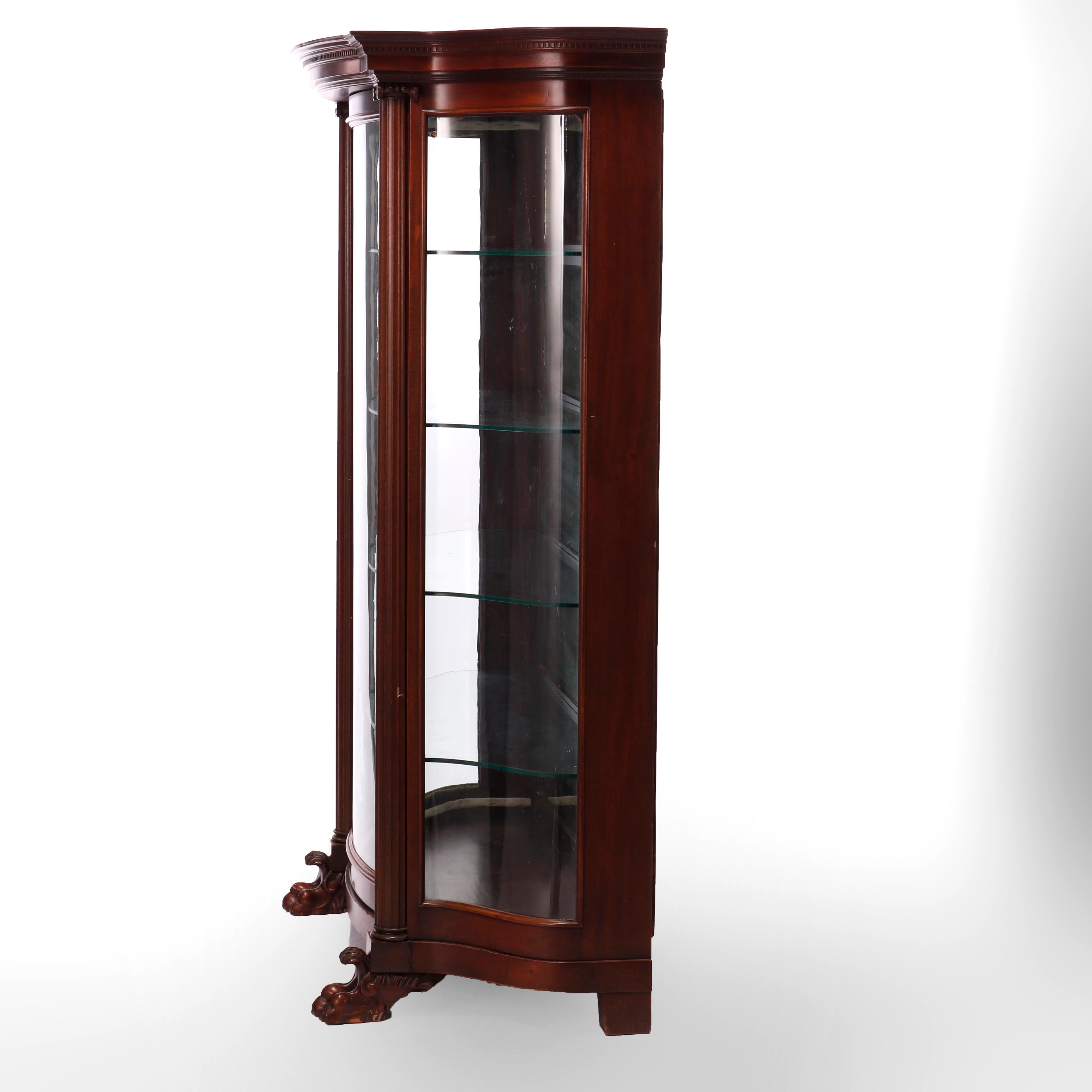 An antique Classical and large china cabinet offers mahogany construction in serpentine form having single curved glass door opening to shelved and mirrored interior flanked by Corinthian column supports and sides also having curved glass, raised on
