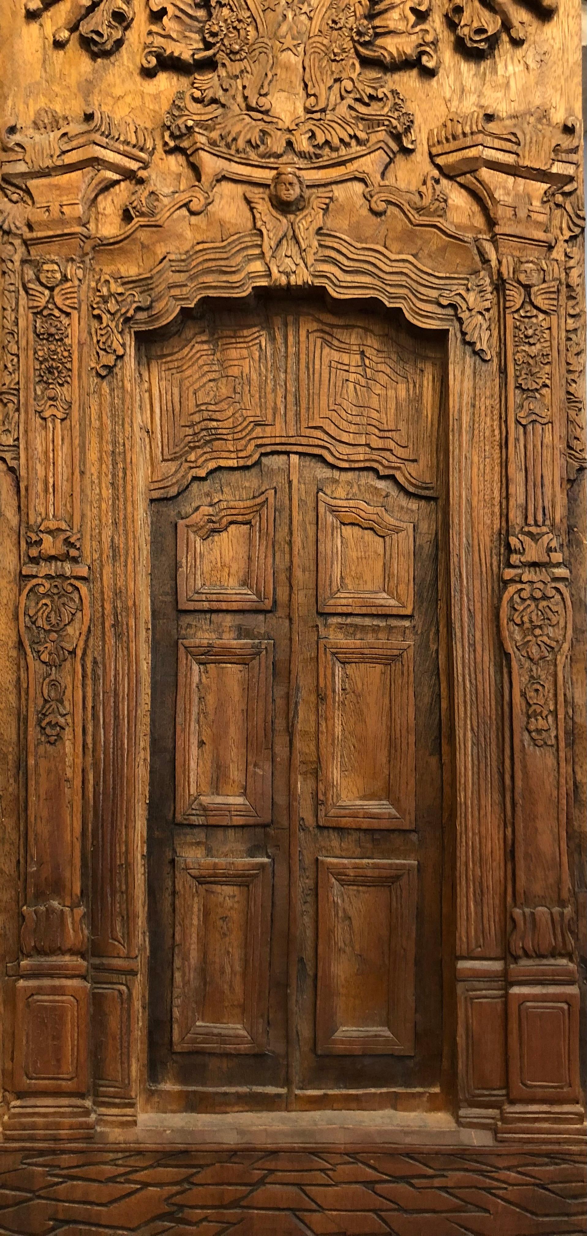 A decorative,  classical carved oak panel with very intricate detail, depicting the facade of a church:  a panelled doorway with fanciful columns on either side,  surmounted by carved angels, scrolls and elaborate pediments. The bottom of the panel