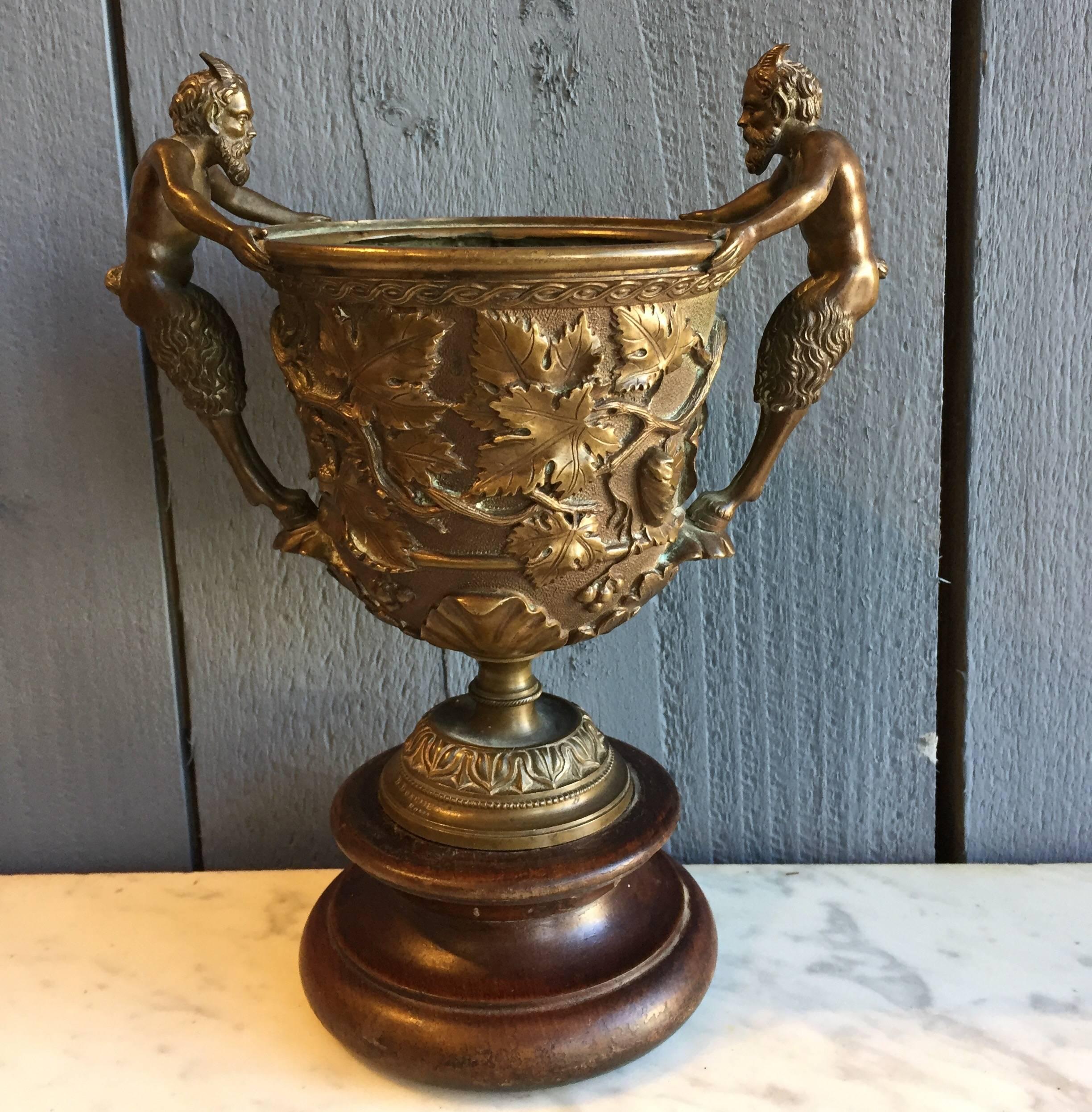 A beautiful Grand Tour patinated bronze cup. Having two nicely executed Faun figure handles. Scrolling leaf and fruit relief to the body and acanthus leaf to the lower part of the vessel. Standing on a turned wooden base. Highly decorative and