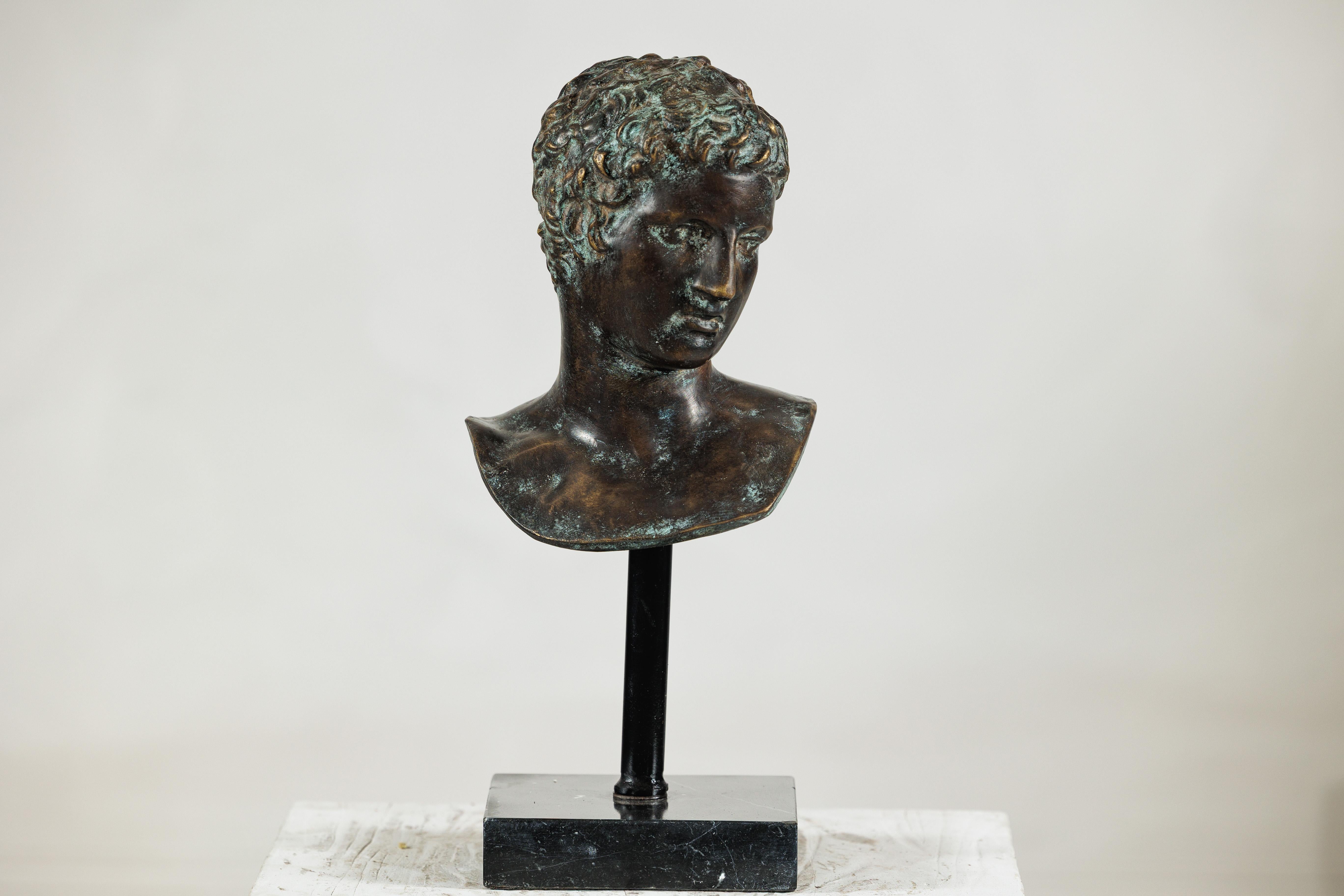 Questo busto in bronzo di un giovane uomo evoca l'eleganza senza tempo della scultura classica. Modellata con riccioli morbidamente dettagliati e un'espressione contemplativa, la testa è presentata su una base di marmo nero che conferisce contrasto
