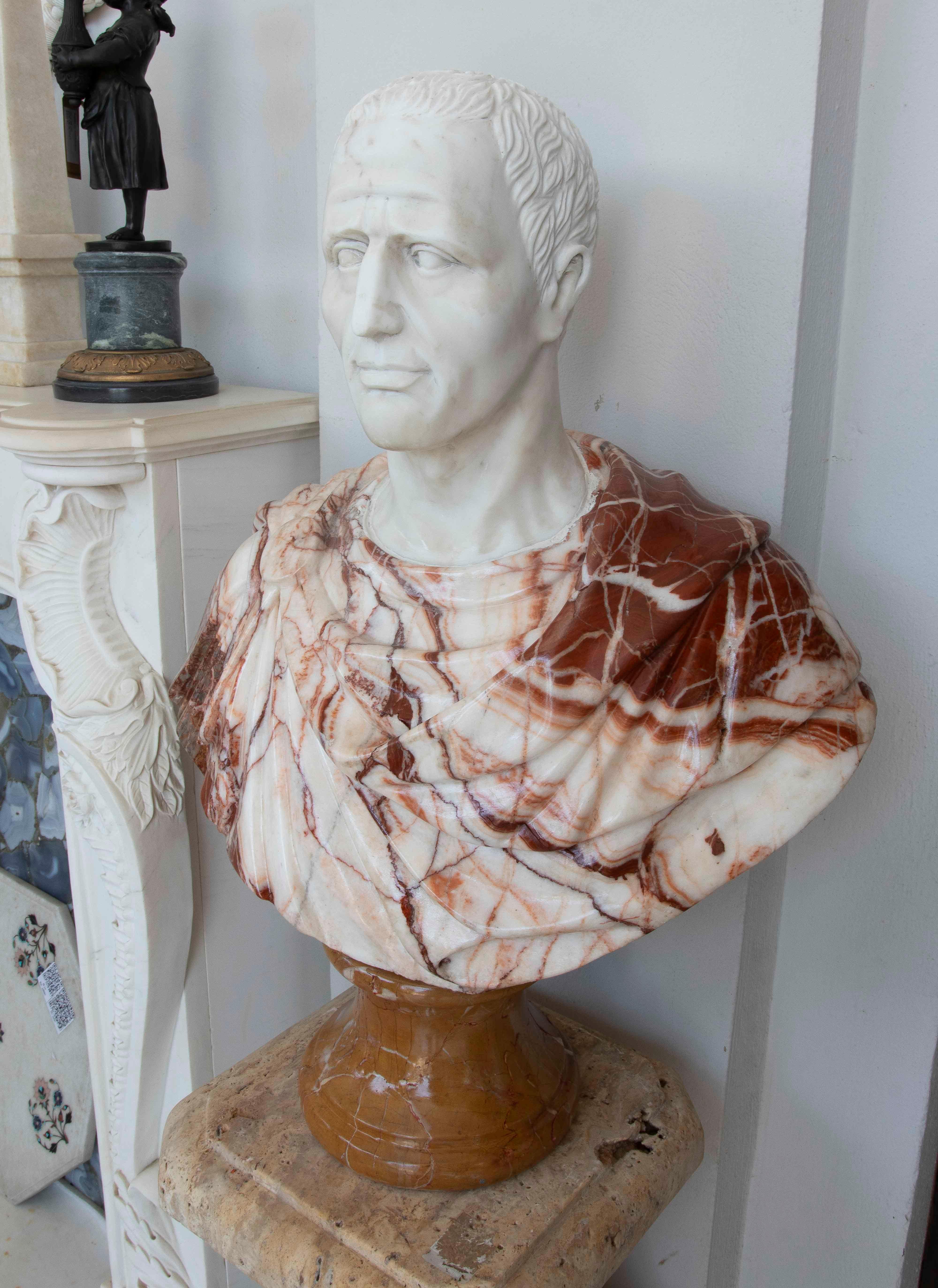Diese imposante Skulptur stellt eine klassische römische Büste dar, deren Gesicht und Hals aus poliertem weißem Marmor und die Toga aus einer spektakulären Kombination aus rotem und weißem Onyx gefertigt sind. Der Kontrast zwischen den natürlichen
