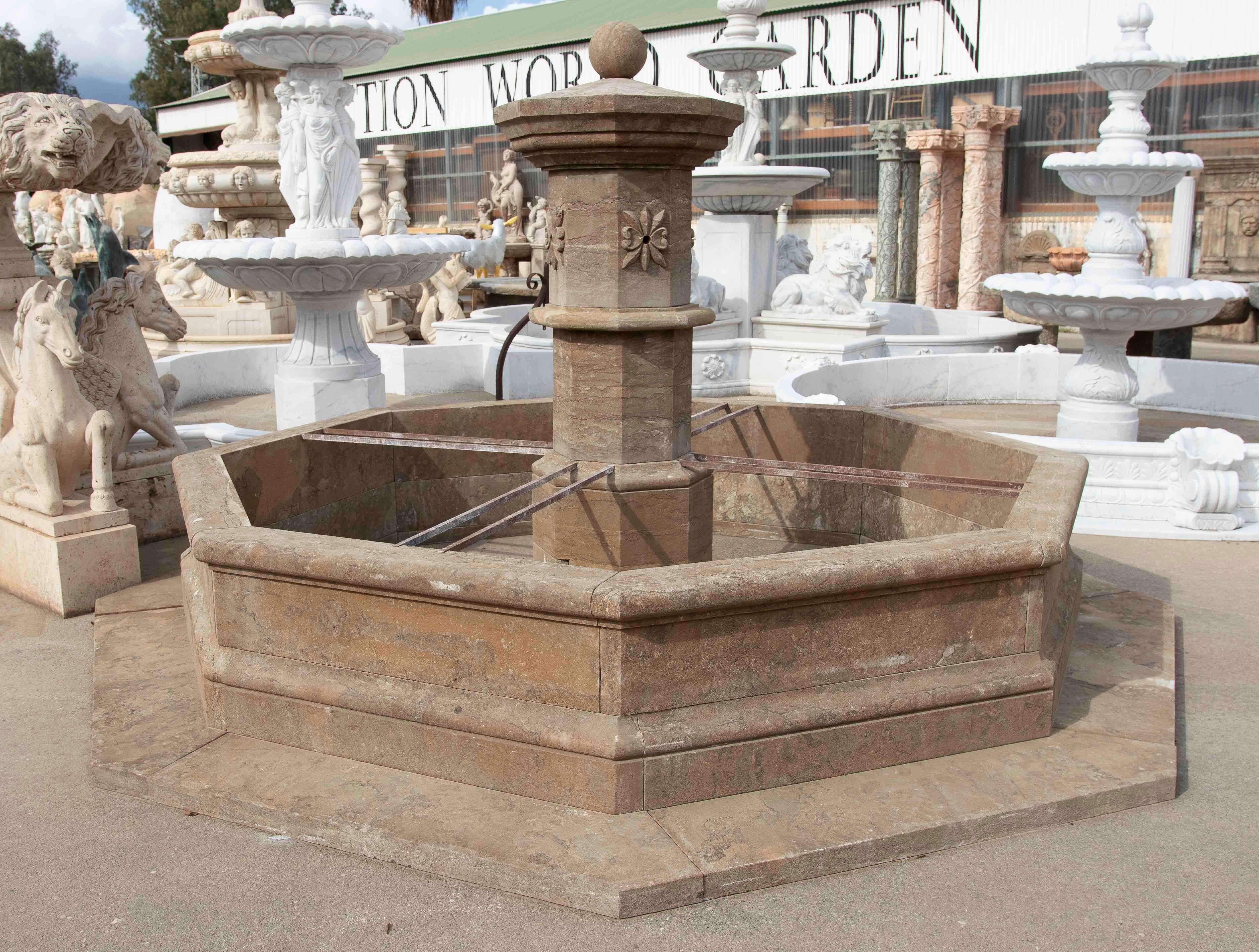 An imposing octagonal fountain carved from warm brown natural stone, featuring an elegant classic design and an ornate top. Its solid structure and balanced proportions reflect traditional craftsmanship, making it ideal for patios, gardens, or