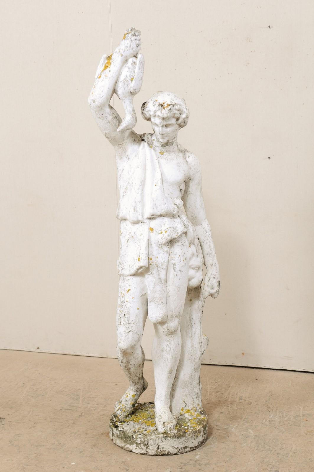 A tall French garden sculpture of male figure from the early 20th century. This antique cast-stone statue from France features a male figure, draped in classical attire, standing against a small tree trunk, with hand raised to display a hanging