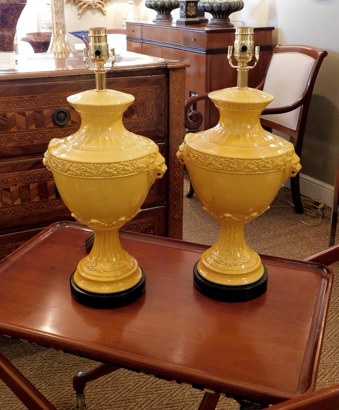 each in a rich egg yolk-yellow glaze with everted neck above an ovoid body with stylized laurel leaf perimeter band flanked by lions masks; all raised on a fluted splayed support resting on an ebonized wooden base.
