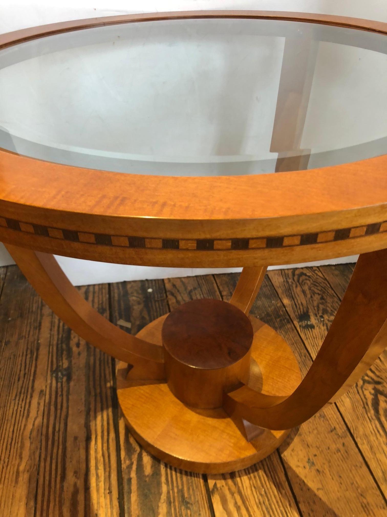 Lovely Art Deco style round end table, a pair is available, having blonde colored high gloss wood with black decorative inlay around the edge and a bevelled glass top. There are 3 curved legs that join at a central cylindrical base at the bottom.