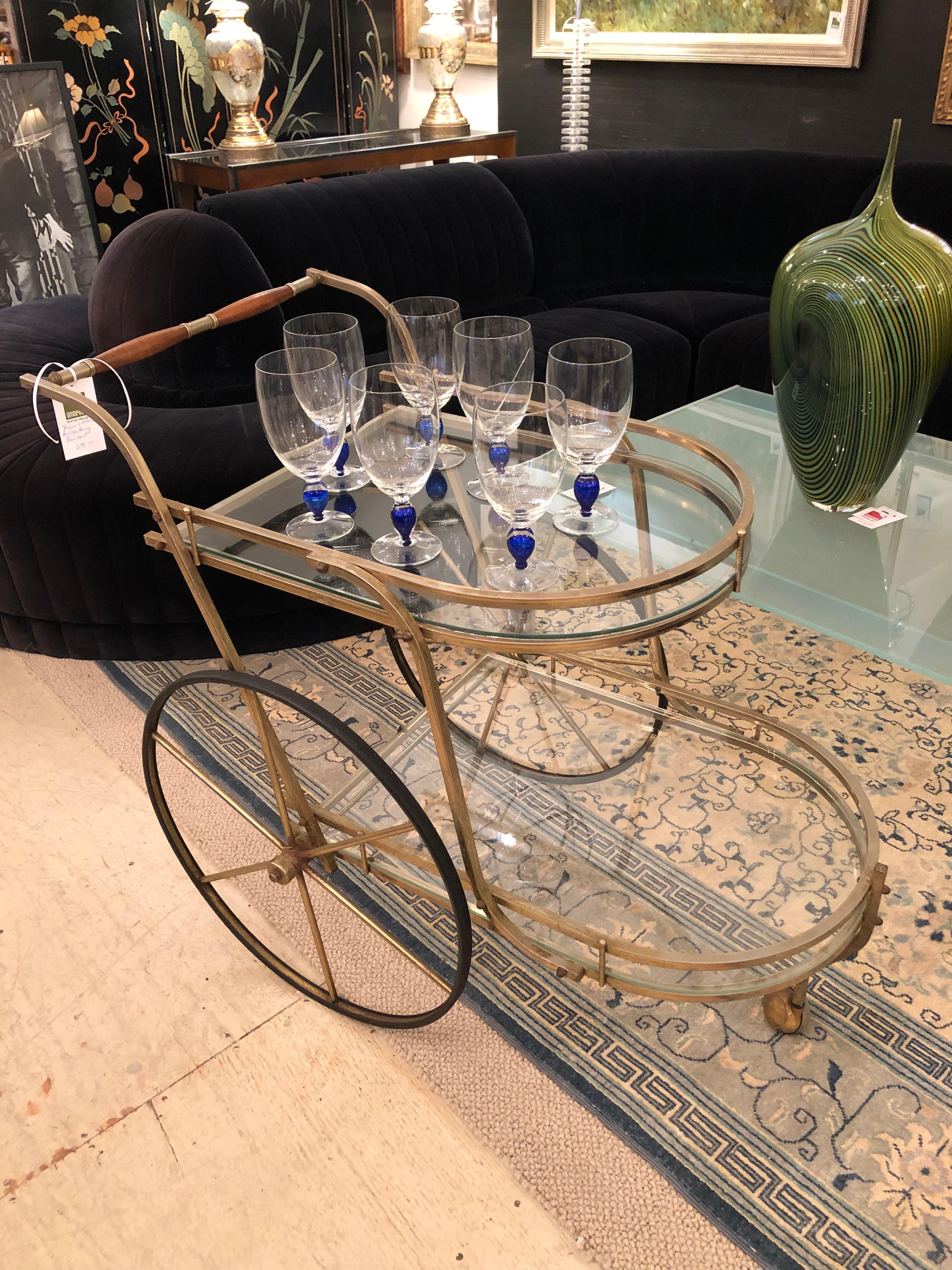 Classy midcentury modern brass and glass bar cart having elegant off set  tiers with curved shape at each end.  The handle is brass and light wood and the large wheels add style to the form.  Rolls easily.  Top tier is 26
