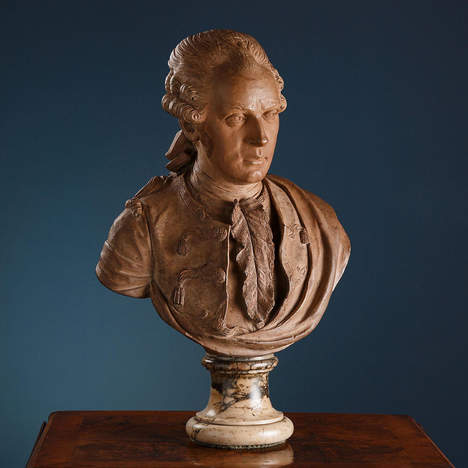The terracotta bust, placed on top of a colored marble pedestal, depicts a male figure with his hair held back in a tail by a ribbon and is attired in a collarless tailcoat closed with frogs with applied tassels and embroidery along the edges, an