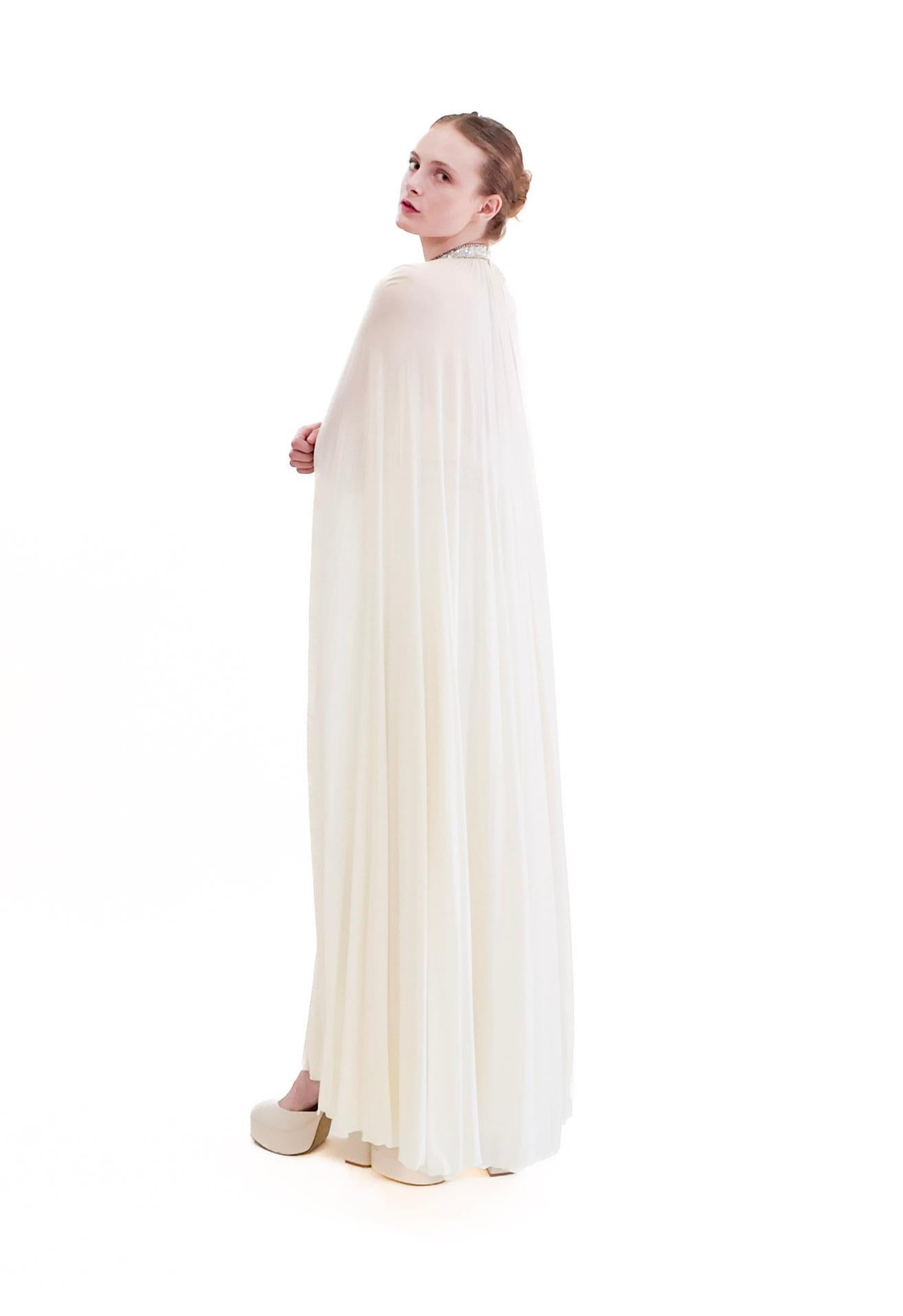 Indulge in sophistication with our Claude Riha white caped gown. Featuring a flowy fabric, this beautiful and elegant piece is perfect for the vintage bride. Adorned with a beaded neckline and waistline, it effortlessly exudes timeless elegance. The
