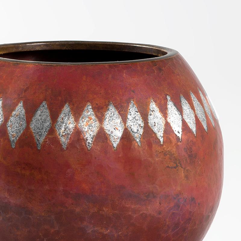 This rare French Art Deco vase, by Claudius Linossier, is a spherical vessel, crafted from hammered copper and brass with geometric silver patina on a burnt umber, red and ochre ground. The powerful red vase features a frieze of diamonds circling