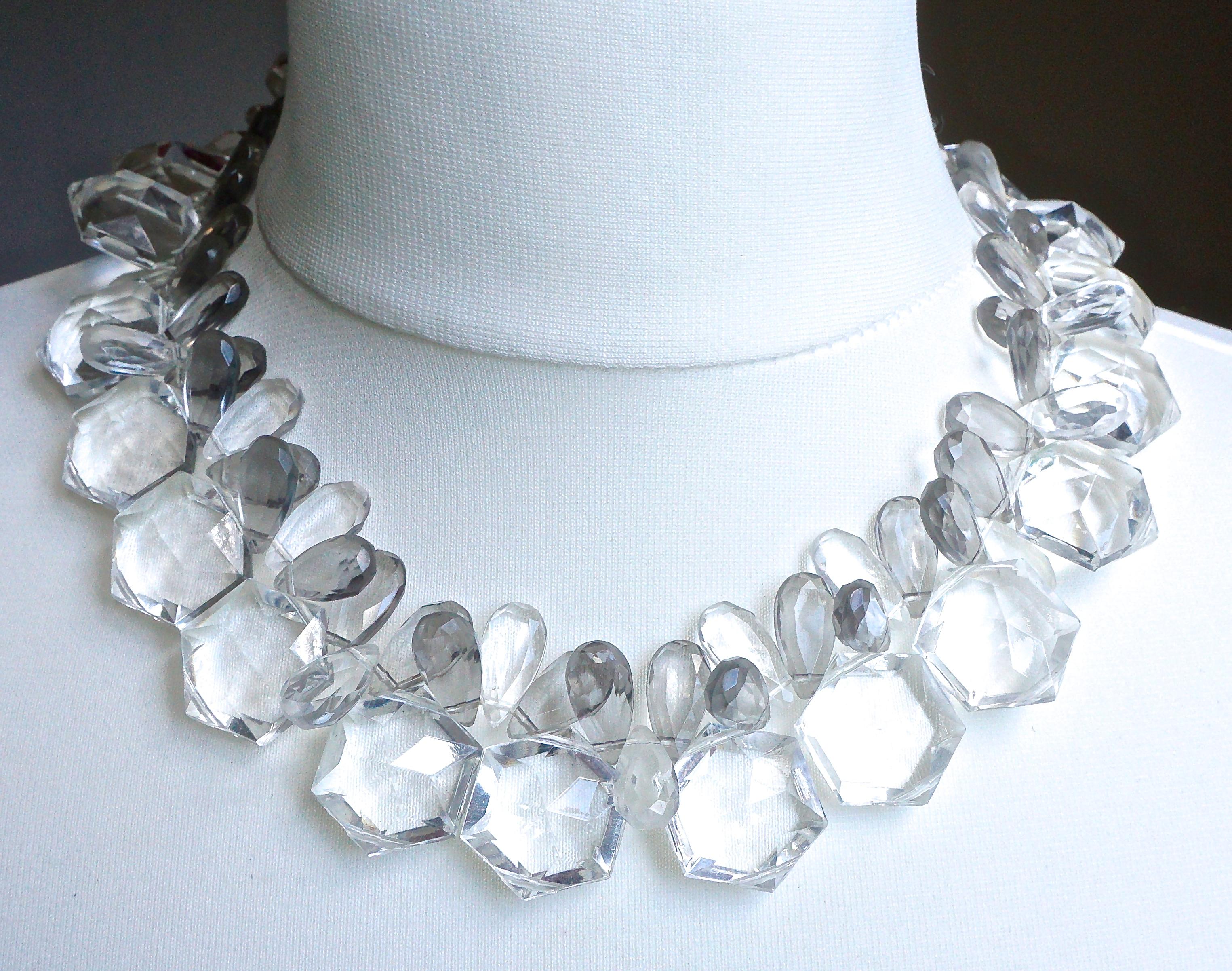 Un joli collier en plastique avec des perles hexagonales transparentes et des perles grises en forme de goutte d'eau, avec un fermoir à pression en argent et perles grises à facettes. Les perles sont enfilées sur un fil blanc. La longueur est de
