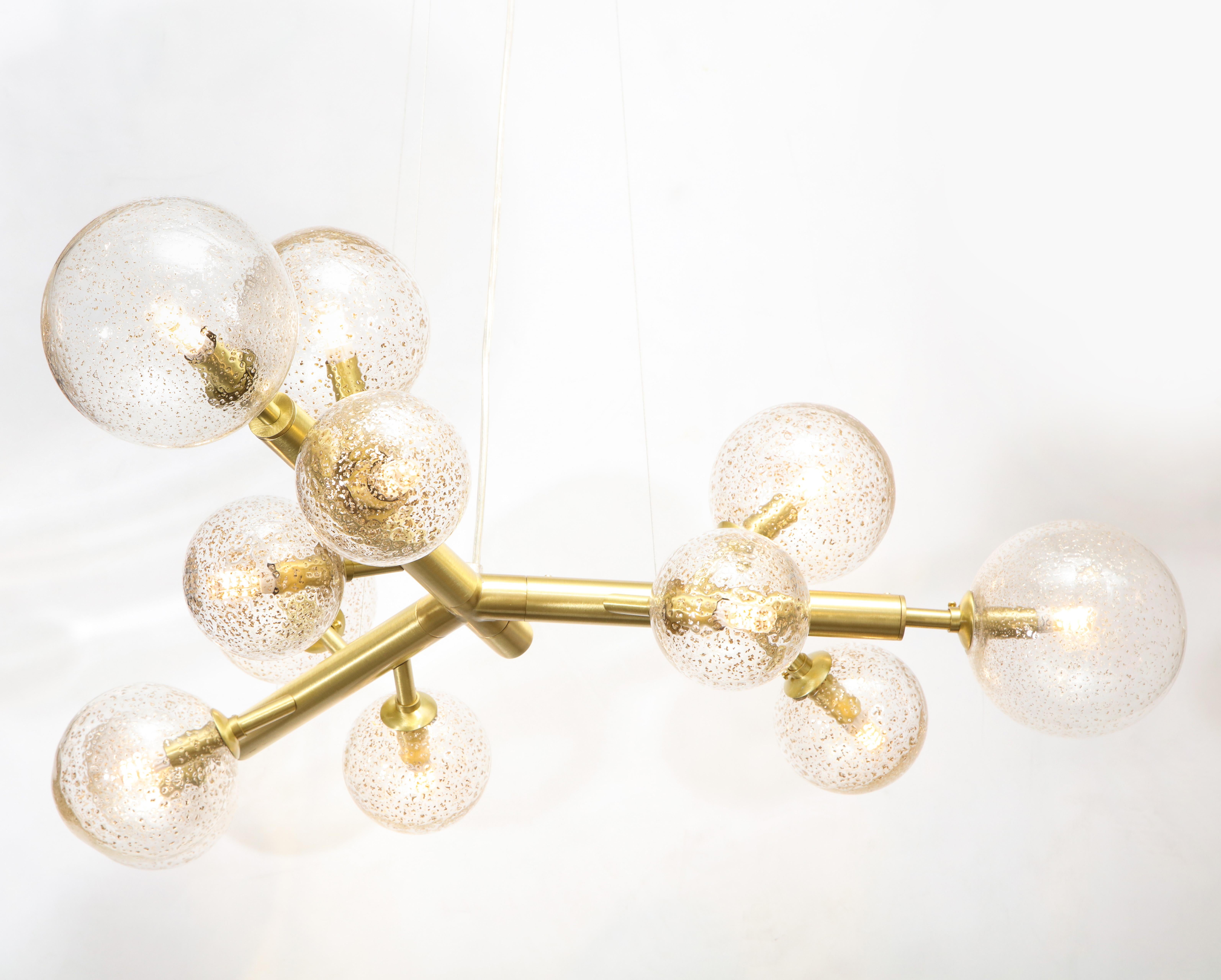 A clear and bronze flecked Murano and brass 12-light globe chandelier by Italian glass maker, Alberto Donà featuring 3 arms each with 4 different hand blown spheres of 3 varying diameters, suspended by clear, adjustable wires that extend to a 6 foot