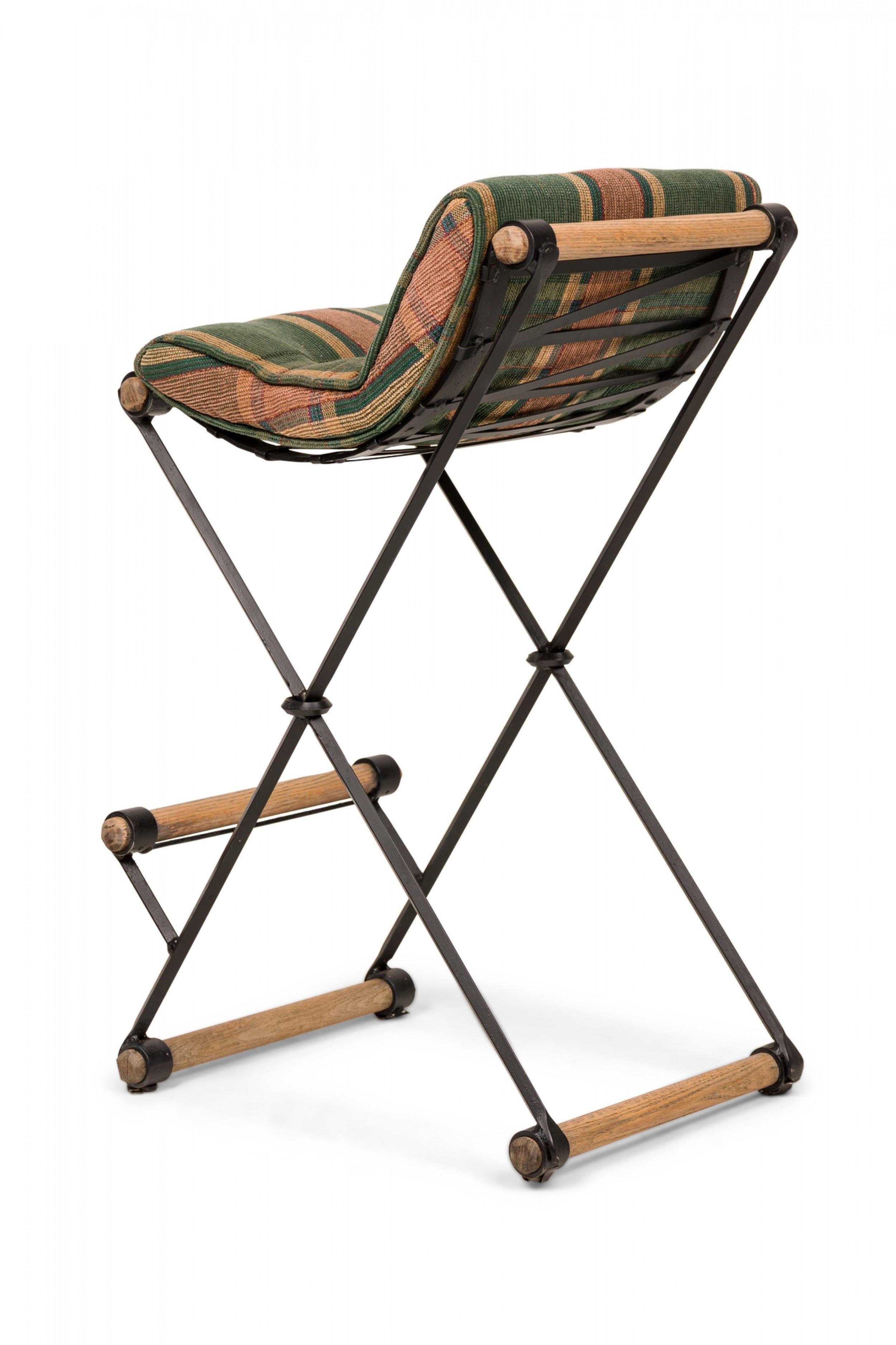 Mid-Century American (1970s) X-frame bar stool with muted green and light brown plaid upholstered seat pad with button tufted detail, resting on a sturdy iron and oak frame. (CLEO BALDON FOR TERRA OF CALIFORNIA)(Other examples in different
