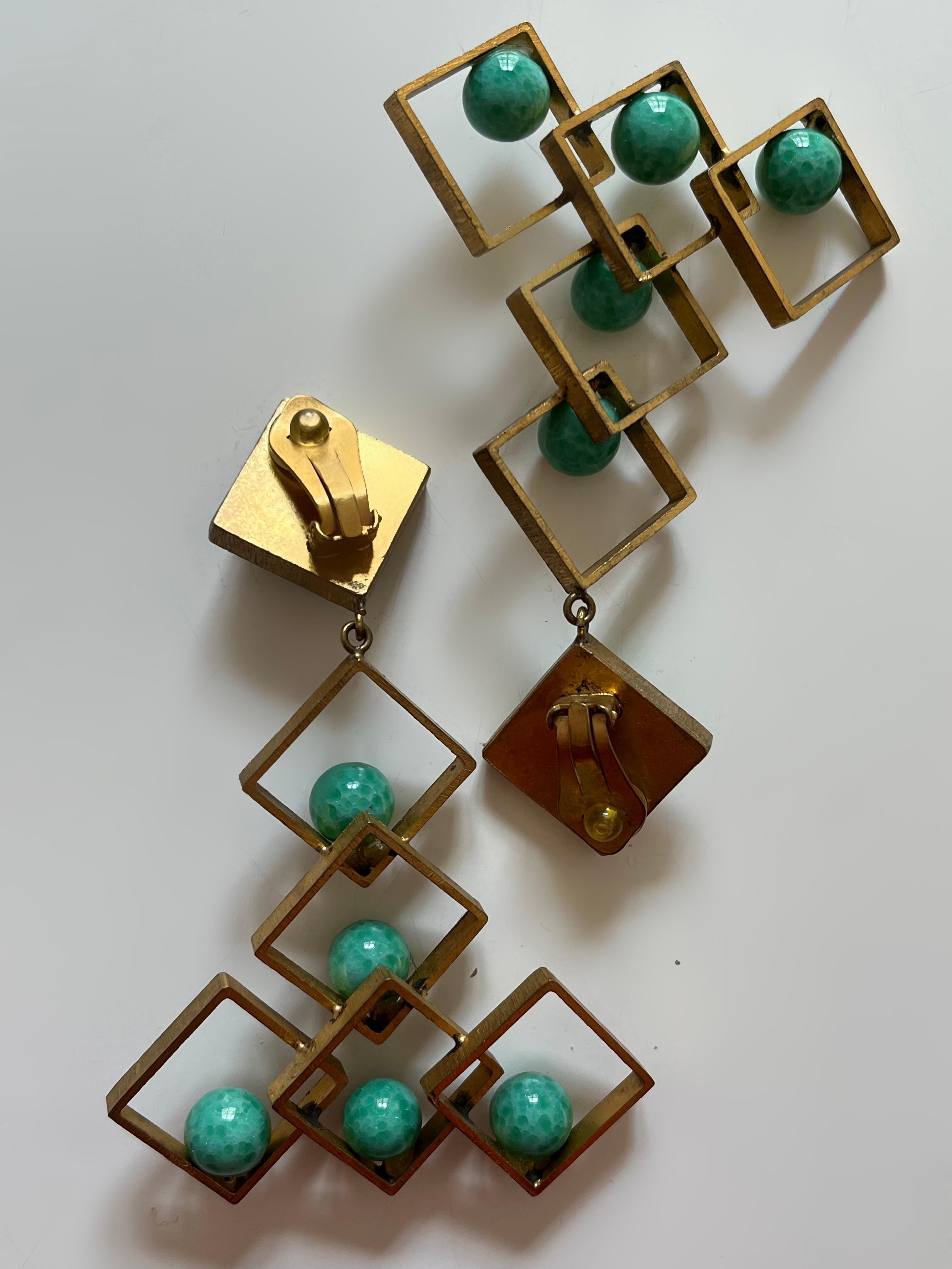 These are stunning geometrical clip-on earrings. Each earring features a cascading arrangement of brass-tone open squares, except for the ones where the clip is attached. Each square is adorned with green slightly marble glass paste beads positioned