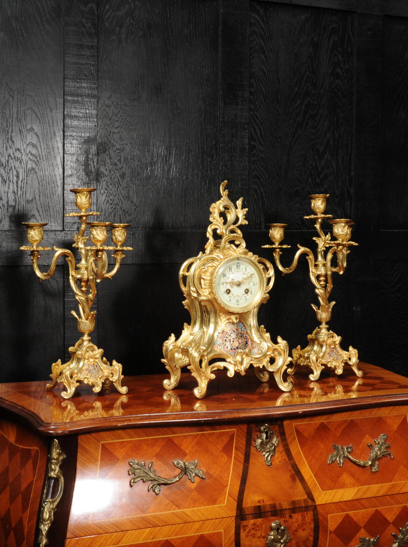 A rare and stunning original antique French clock set, in gilt bronze. It is beautifully designed in the Rococo style and features exquisite cloisonné or champlevé enamel work to both the clock and candelabrum. It has an elegant waisted, asymmetric