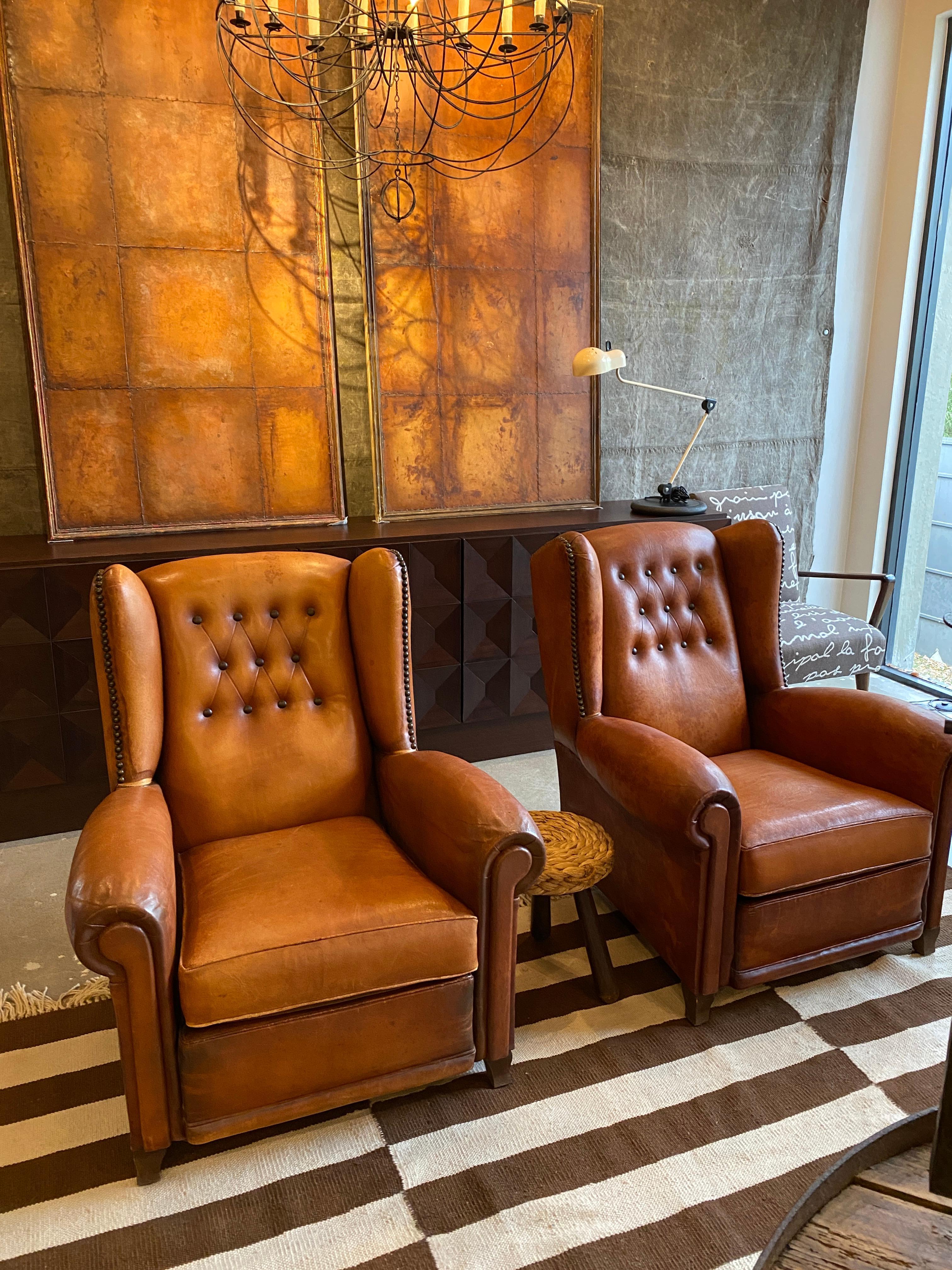 Pair of cognac leather wingback chairs with tufted backs and large nailheads. Nicely matched pair in good condition. Leather has luster, patina and shows some repair. Unusually comfortable and generous scale in comparison to many of the same period.
