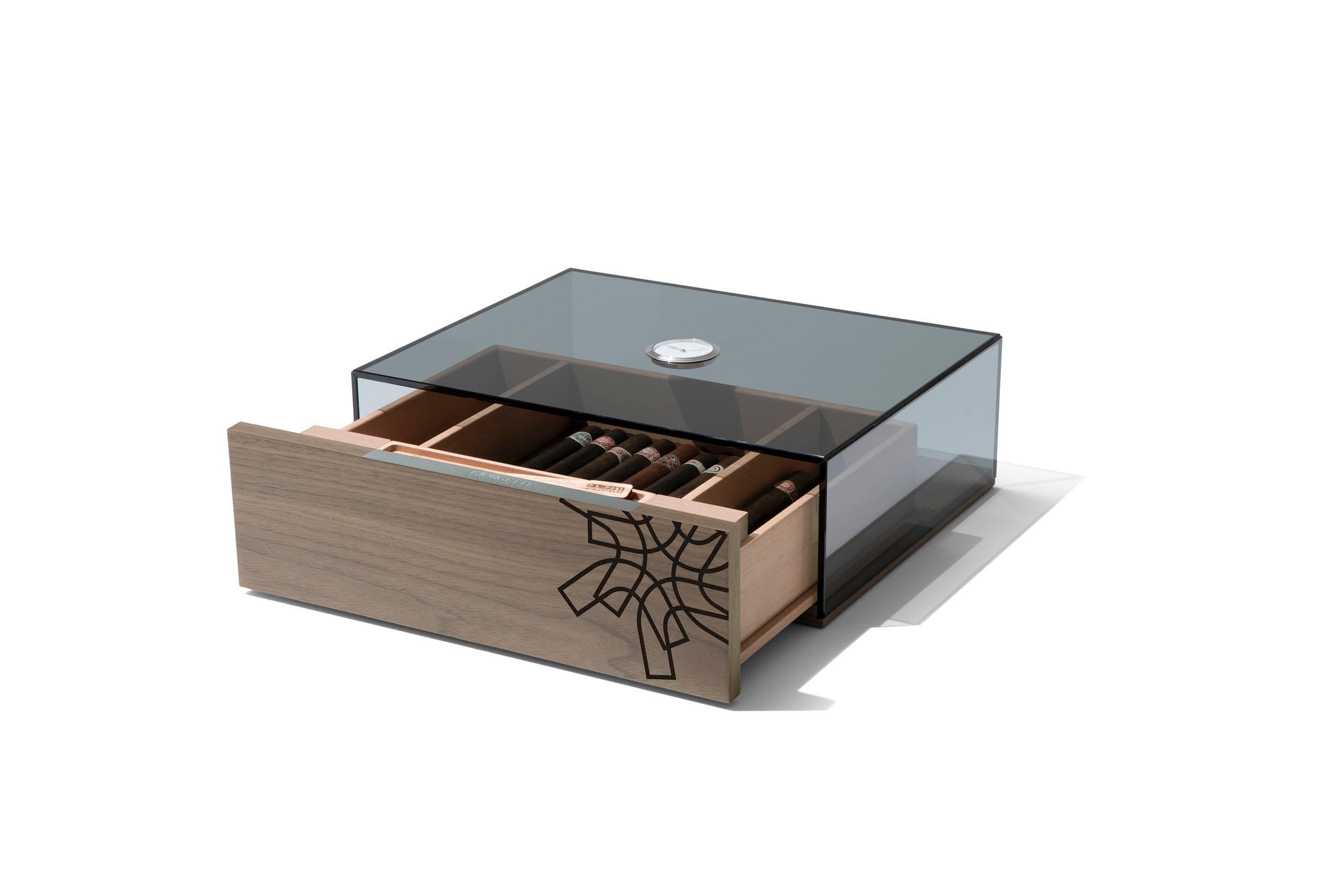 Caja para puros en acabado gris nogal canaletto (fin. 2W) con pictograma incrustado en la parte delantera en wengé. Interior y tabiques de cedro español natural sin tratar.
Tapa de cristal gris con higrómetro analógico integrado. Humidificador