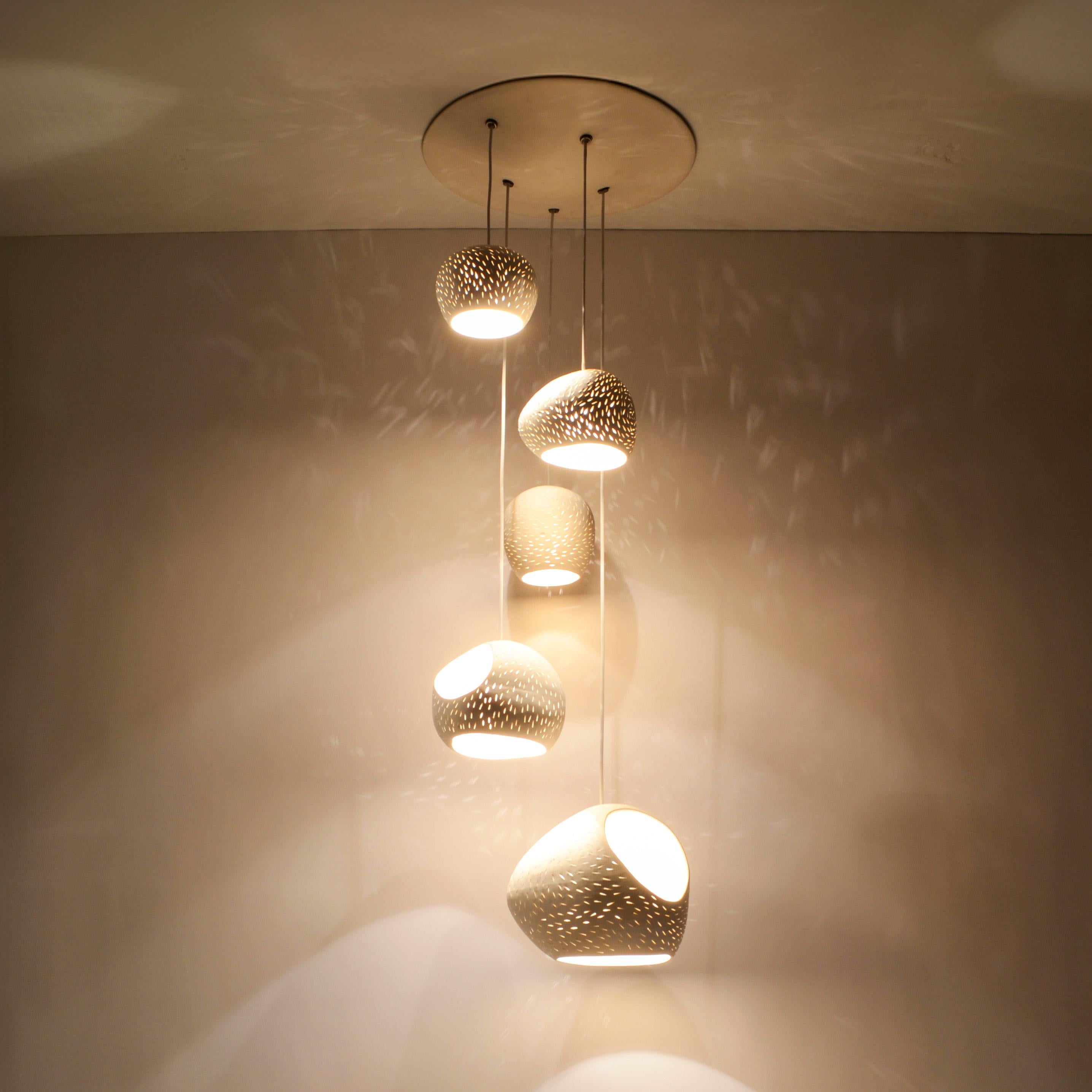 Ce lustre suspendu de designer est une véritable sculpture de l'éclairage. Le design fonctionnel présente cinq nuances de céramique blanc chaud de formes différentes, créant un jeu captivant de silhouettes symétriques et asymétriques. Ce lustre