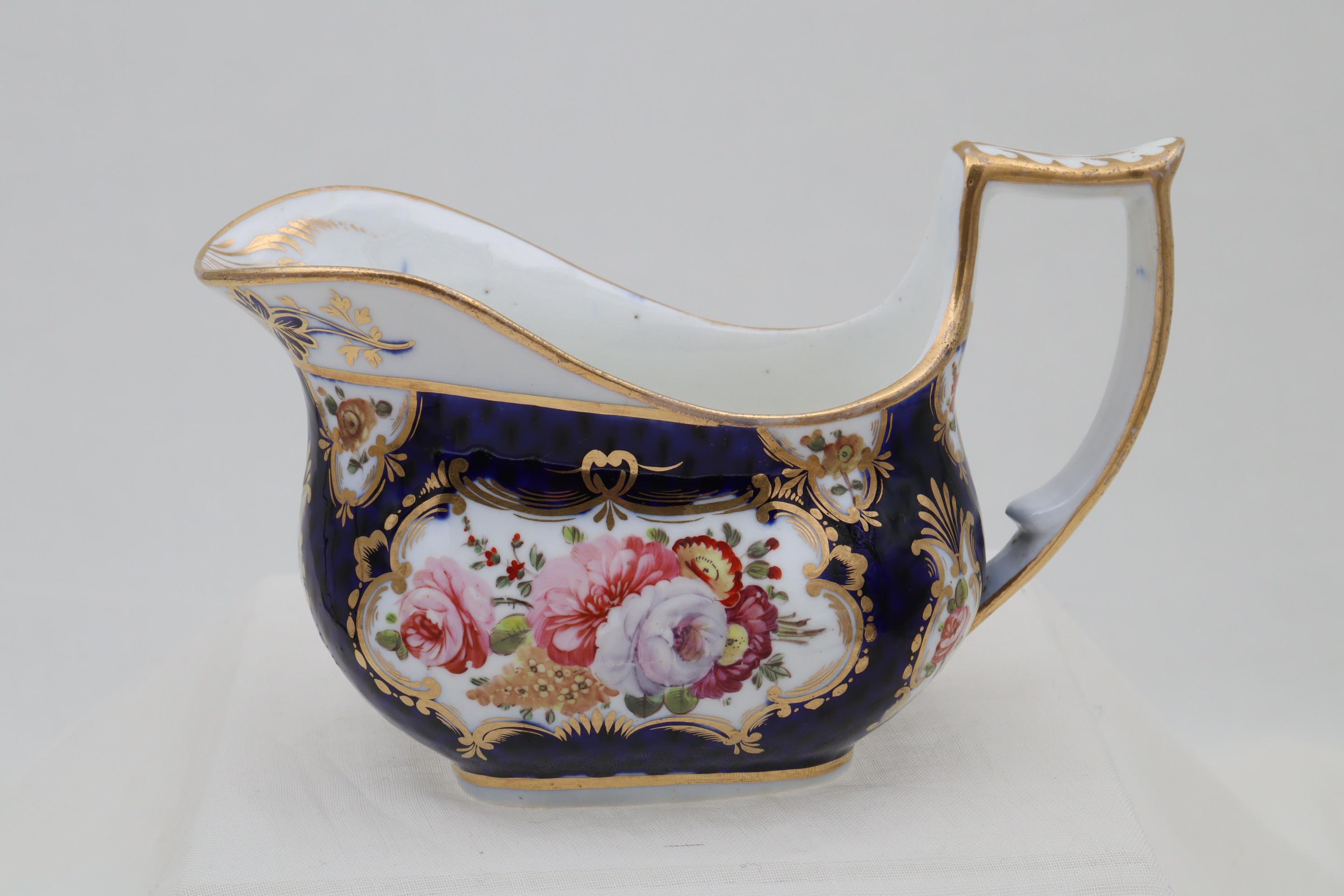 This very attractive hand painted porcelain creamer is by John Rose Coalport and is decorated with cartouches of colourful floral sprays, outlined in gilding, all on a Worcester type scale blue ground. The shape of the creamer is known as the London