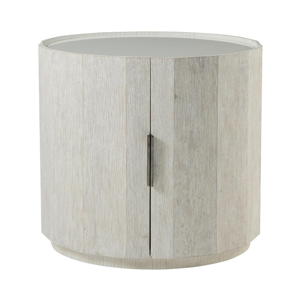 In our Sea Salt Finish, this side table crafted from wire-brushed cerused pine displays a faceted circular exterior of mitered panels with a white limestone top. With two doors with organic ribbed metal hardware that lead to additional storage