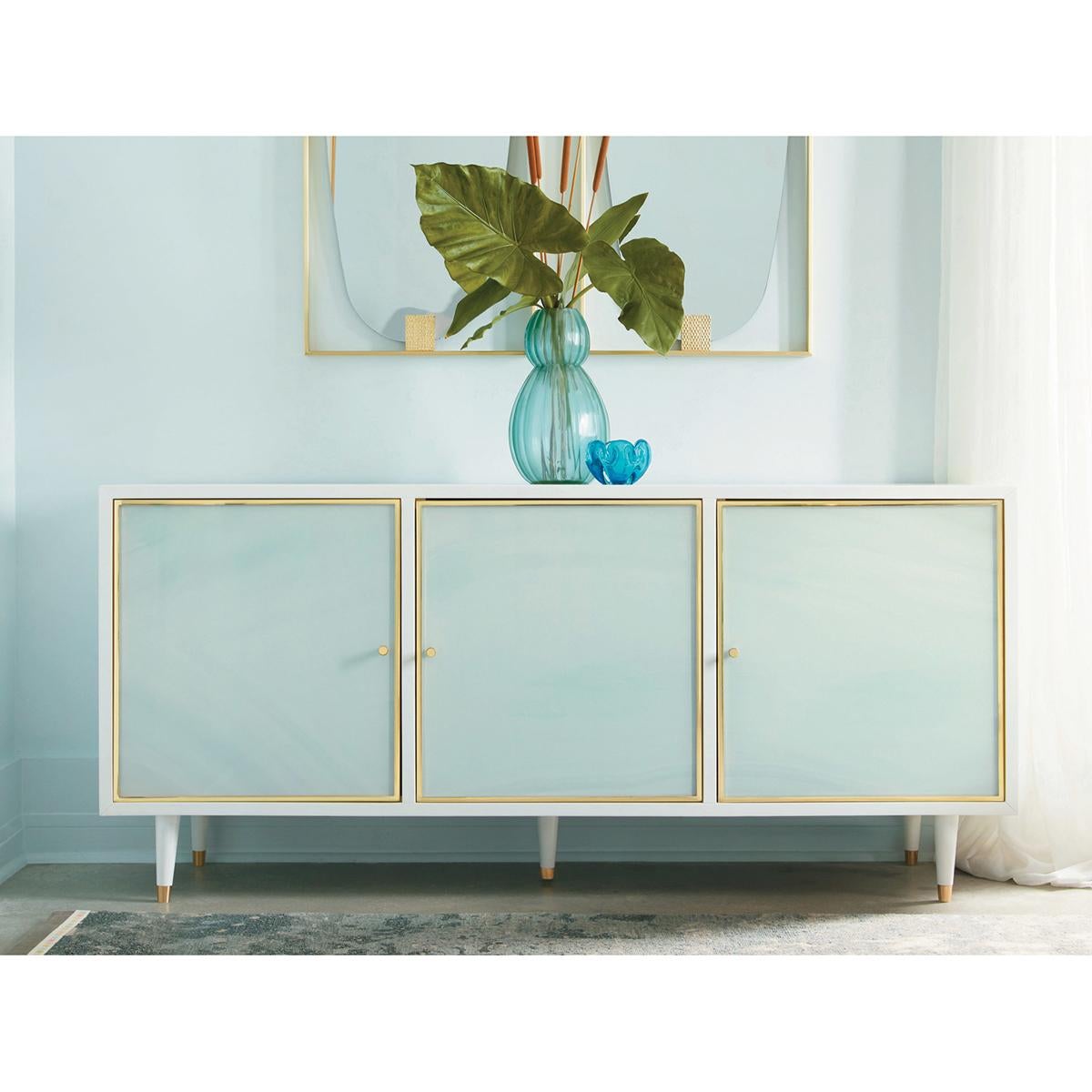 Coastal Sea Glass Credenza. With a white lacquer case and sea glass acrylic door fronts, this three-door cabinet has brass trim framed doors and knob pulls. Raised on turned and tapered legs.

Dimensions: 70