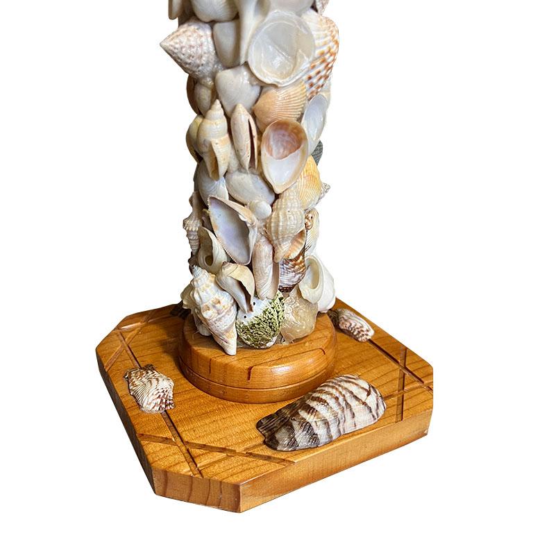 A tall wooden lamp encrusted with natural seashells. This piece will be a wonderful addition to a living room or nightstand. The lamp itself is created from wood and has a square base with carved detail. The body of the lamp is encrusted with