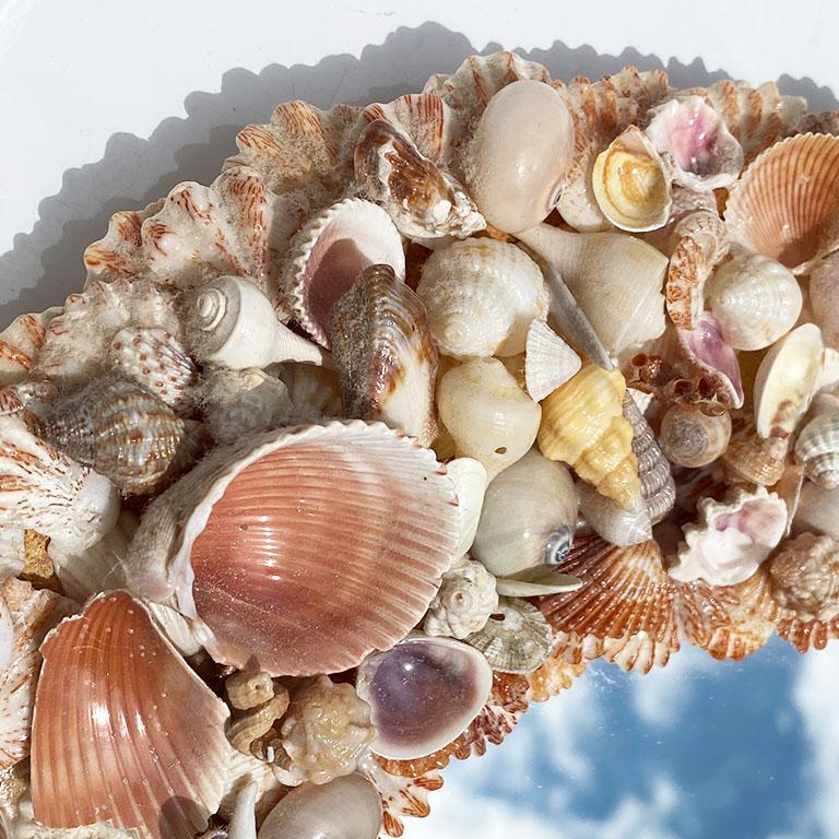 A shell encrusted mirror that will bring the beach indoors. A perfect reminder of the beach, this mirror is oval in form and created from wood. Shells are painstakingly placed throughout the front and border of the mirror to create the coastal look