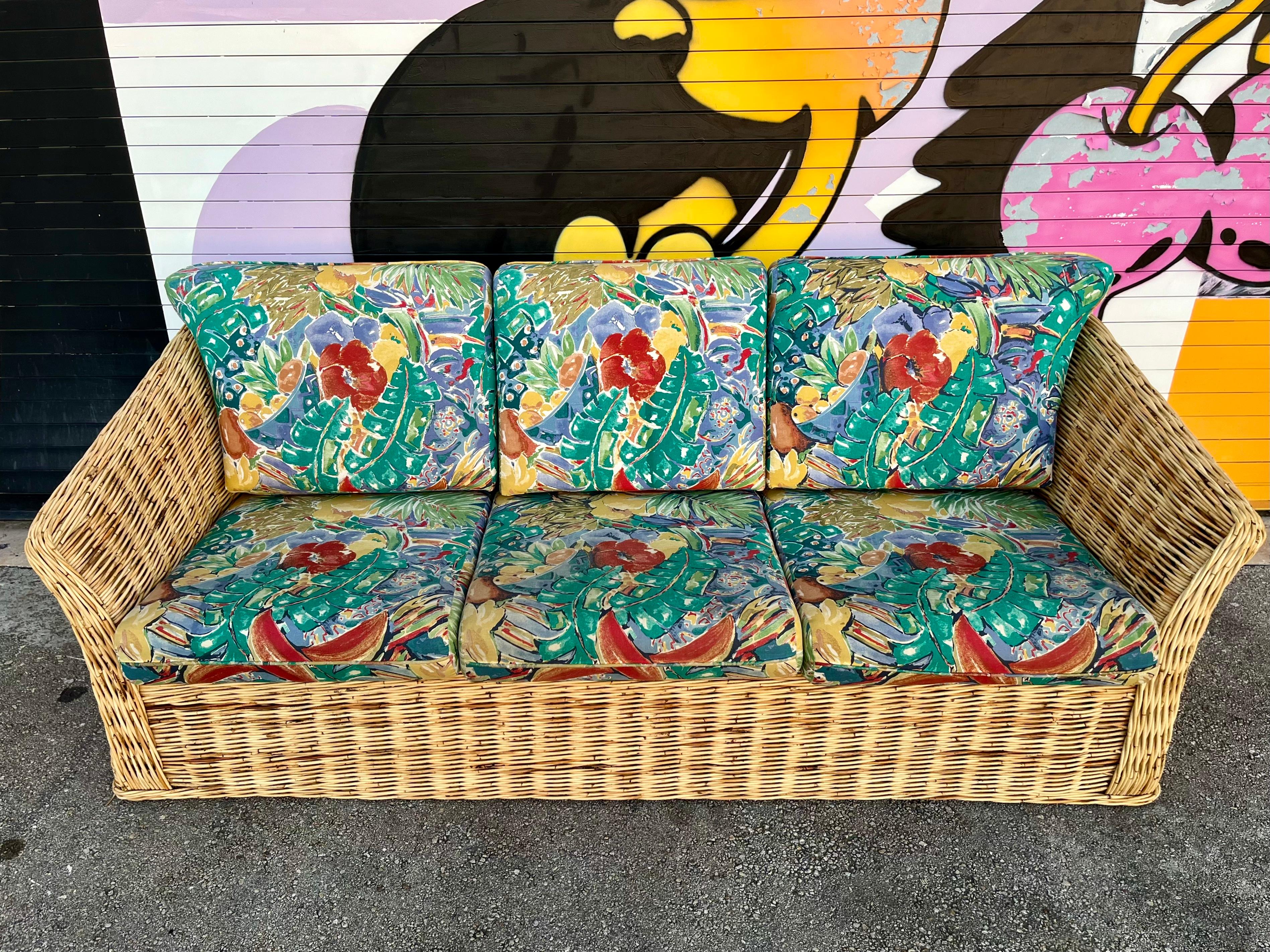 Vintage Coastal Style / Bohemian Woven Wicker Three Seat Sofa. Circa 1980s 
Features a gorgeous sculpted wicker body throughout the entire sofa, with removable cushions with the original 1980s tropical print textile upholstery. 
In excellent