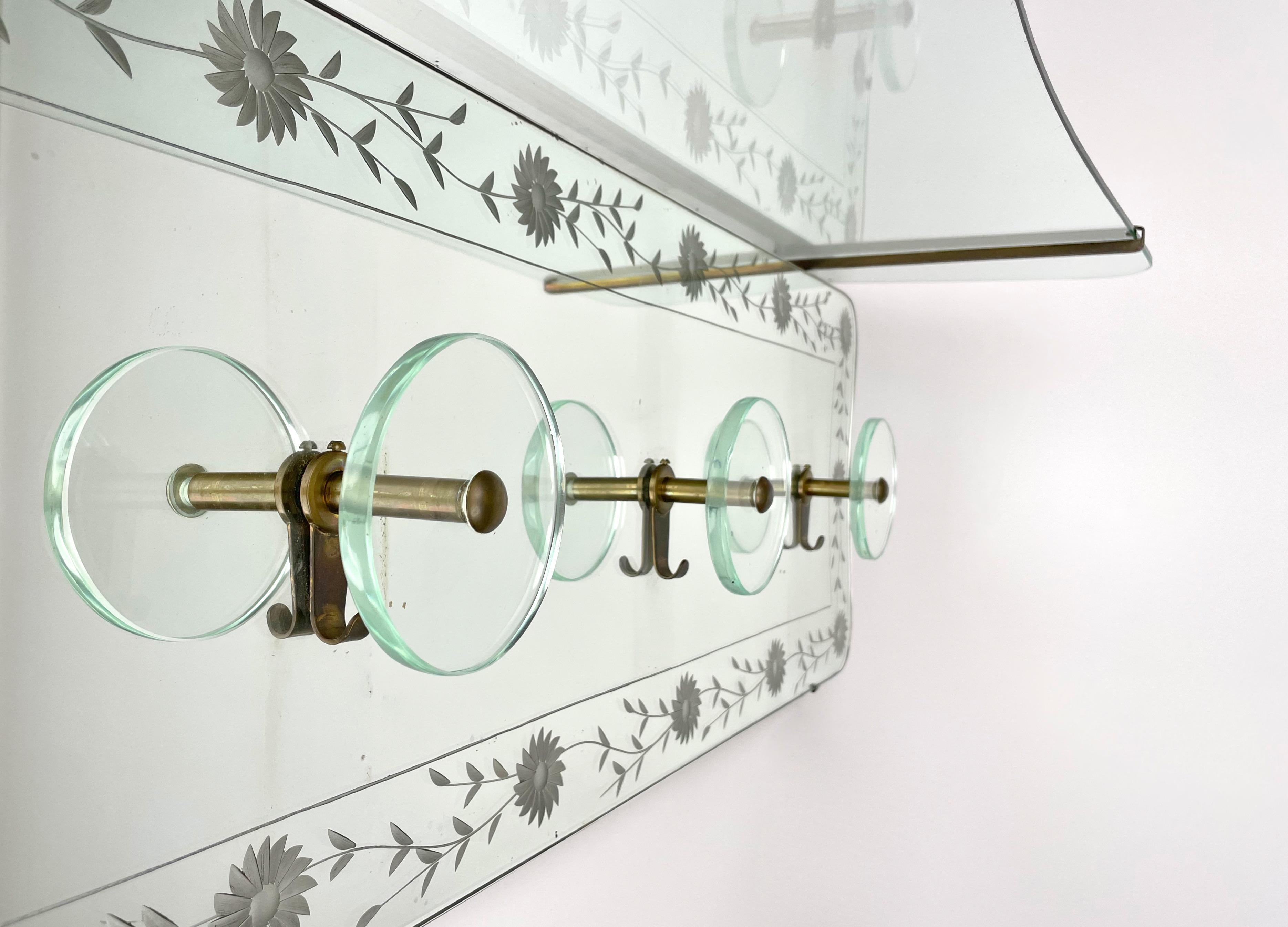Wall-mounted coat rack in mirror with a top glass shelf supported by brass strucure featuring elegant carved details and four circular hooks in glass and brass by Cristal Art.

Made in Italy in the 1950s.