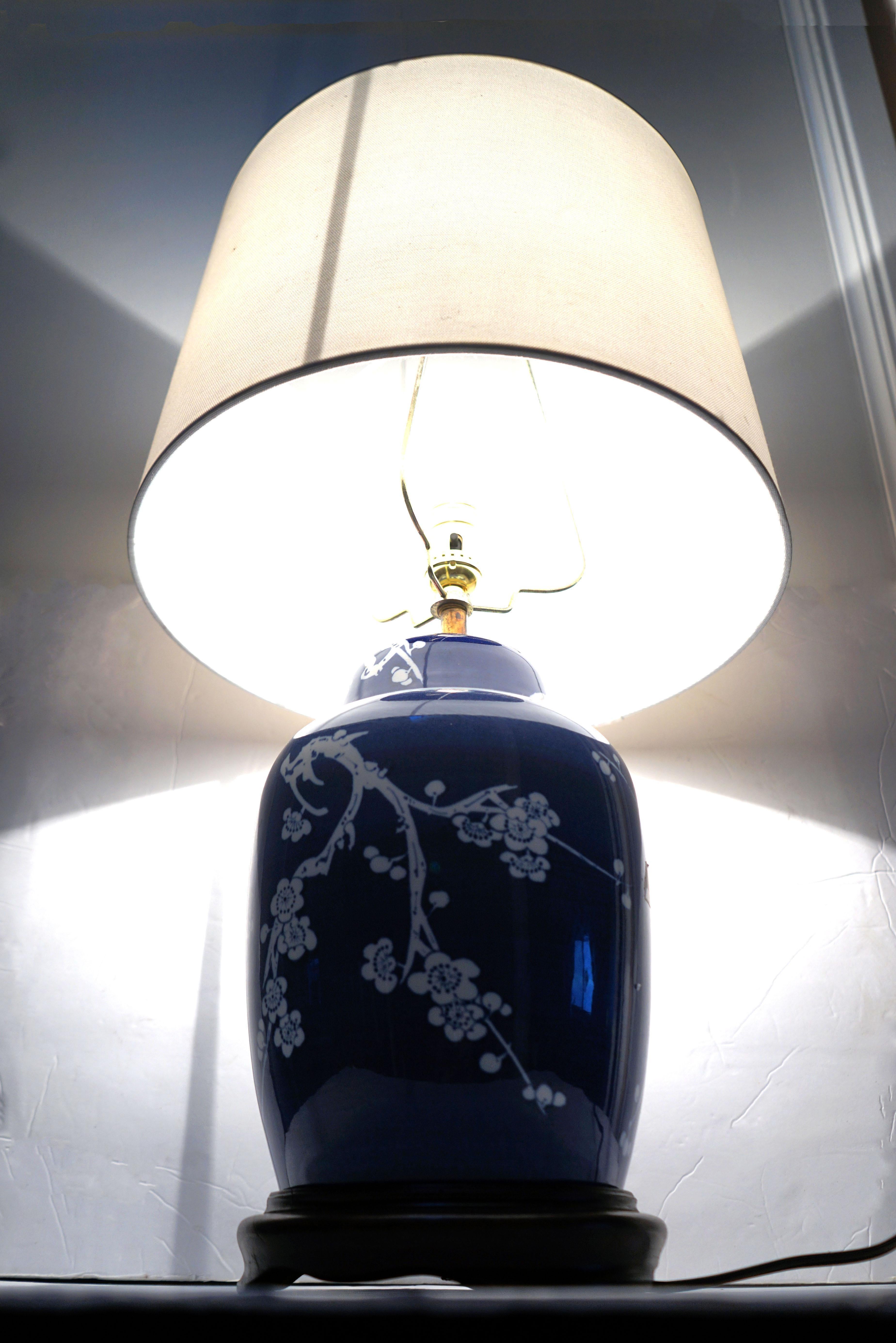 The blue cobalt and white contrasting prunus or cherry blossoms on a ginger jar lamp are exceptional. The blue and white Chinese ginger jar lamp is delightful. The contrast is sharp on a walnut base with a round shade that is original to the piece.