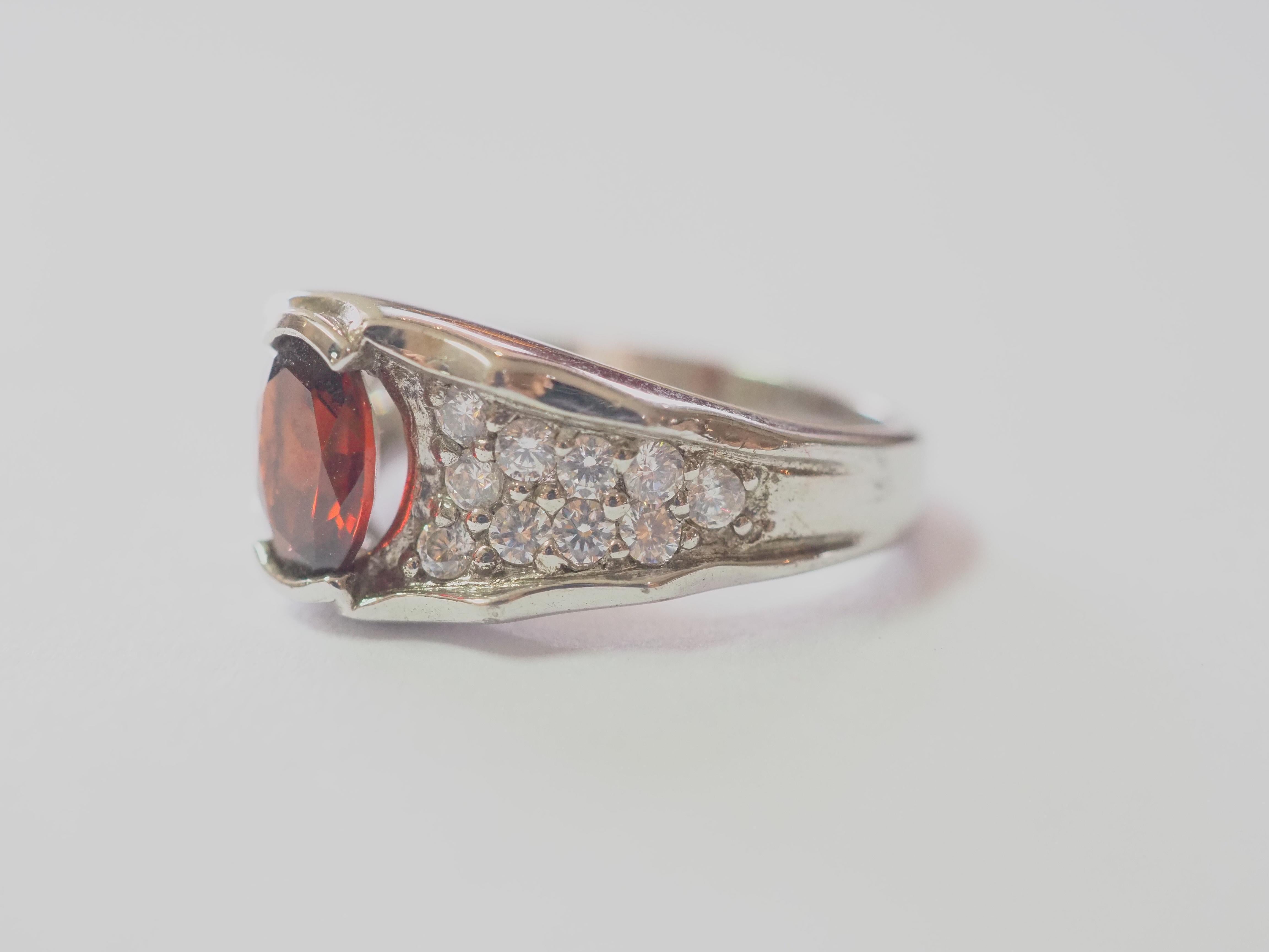 This ring is a beautiful and elegant cocktail ring in solid sterling silver. The ring is decorated by oval garnet channel set in the middle. The brilliant white stones surrounding the red gem are cubic zirconia. The garnet is very widely popular due