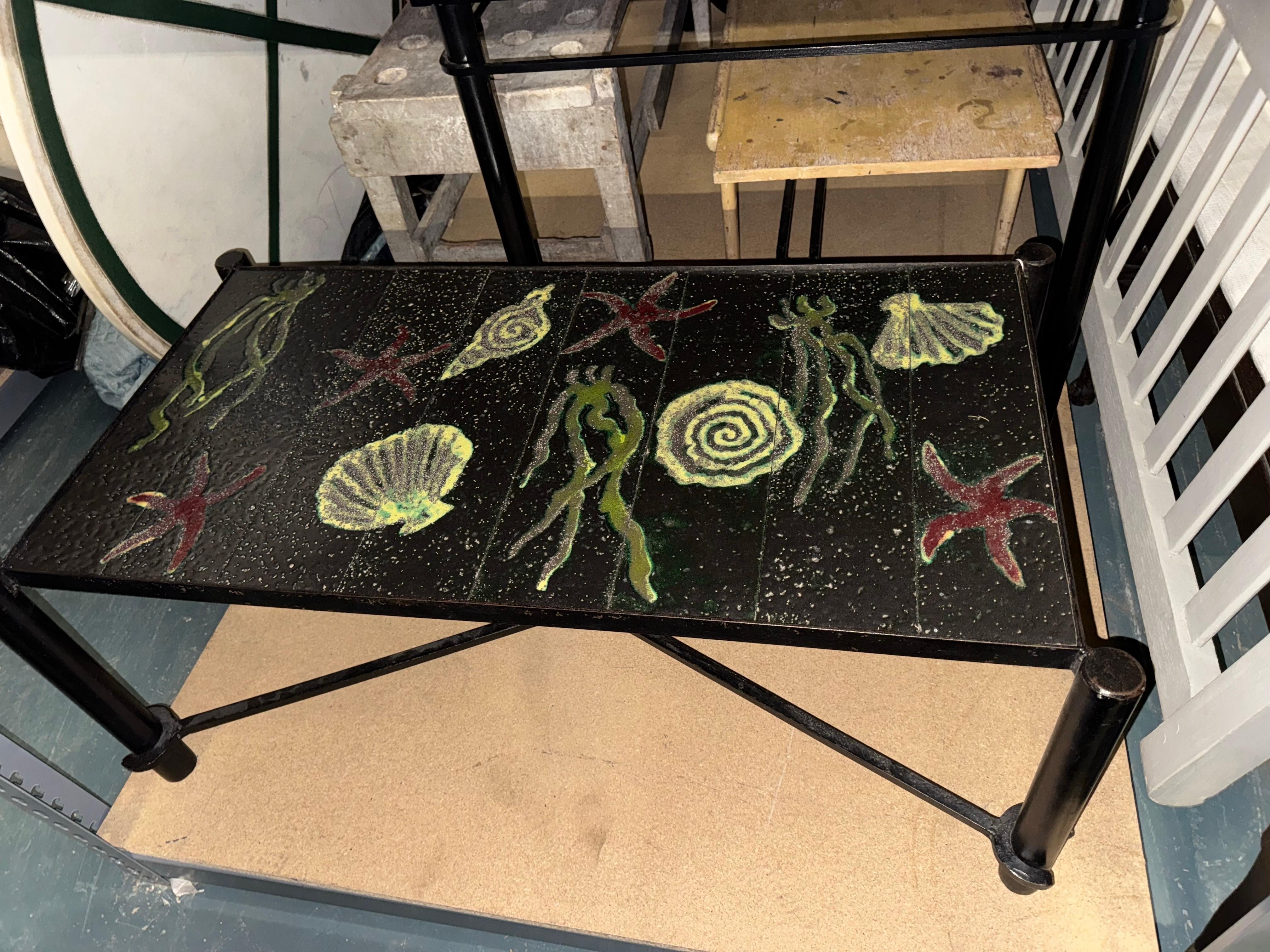 An occasional table attributed to Jacques Adnet, featuring a rectangular top decorated with a series of stylized marine forms executed in vivid green and red pigment. Imagery of shells, starfish, and abstract aquatic figures is set against a dark,