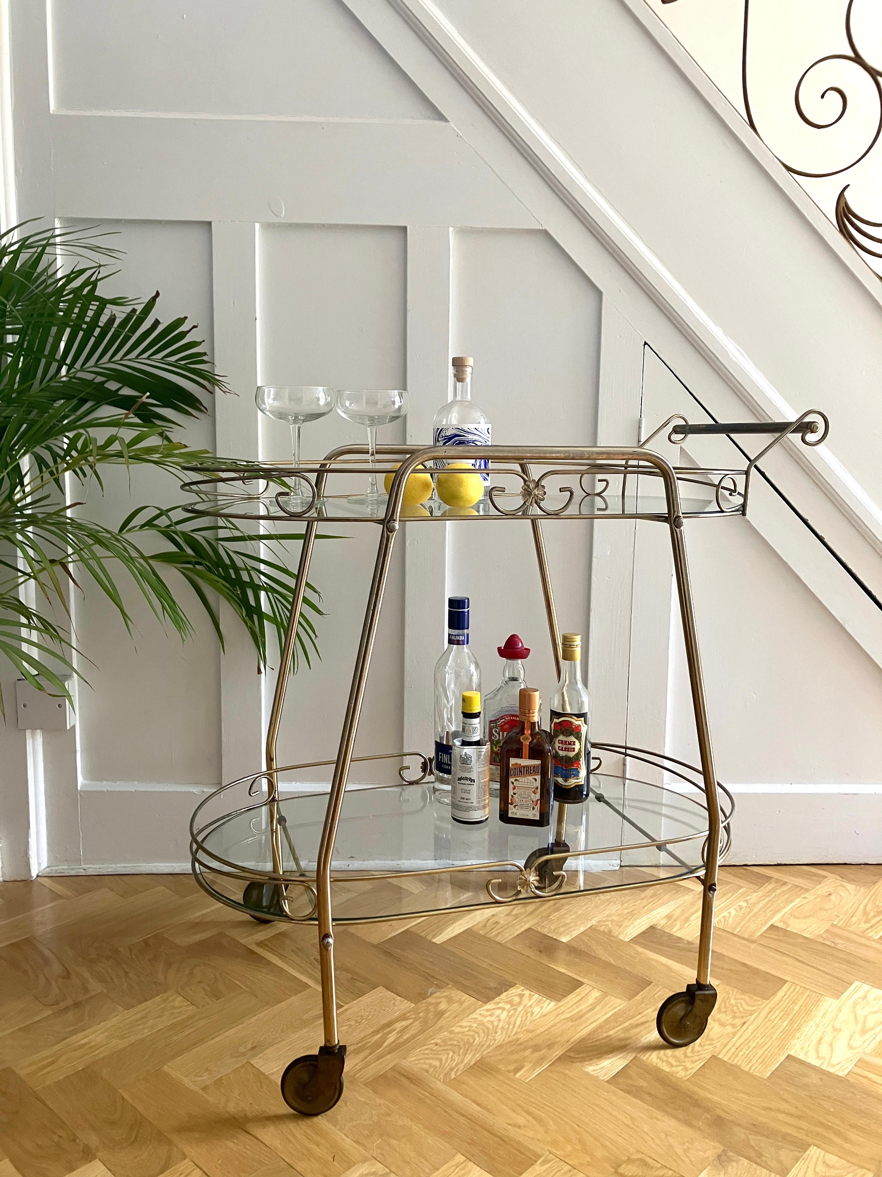 A gorgeous mid-century cocktail trolley with a brass-plated finish. The drinks cart has attractive metal swirl decorations and small flower motifs on the sides. There is a push handle and two sidebars for lifting the trolley.
The drinks trolley has
