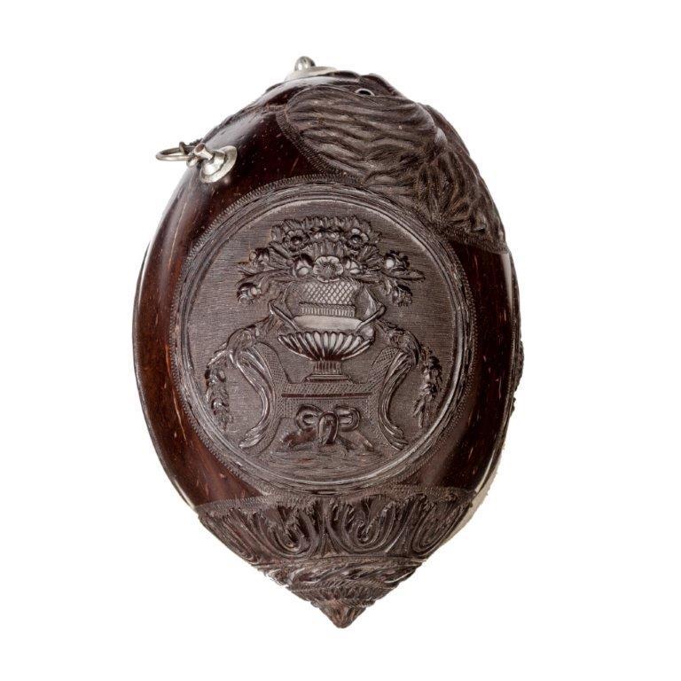 A very fine coconut shell “bugbear” powder flask with silver mounts, the top finely carved with a lion’s mask with rippling mane and glass eyes, the central frieze with ovals depicting musical instruments, a floral arrangement and a basket of fruit,