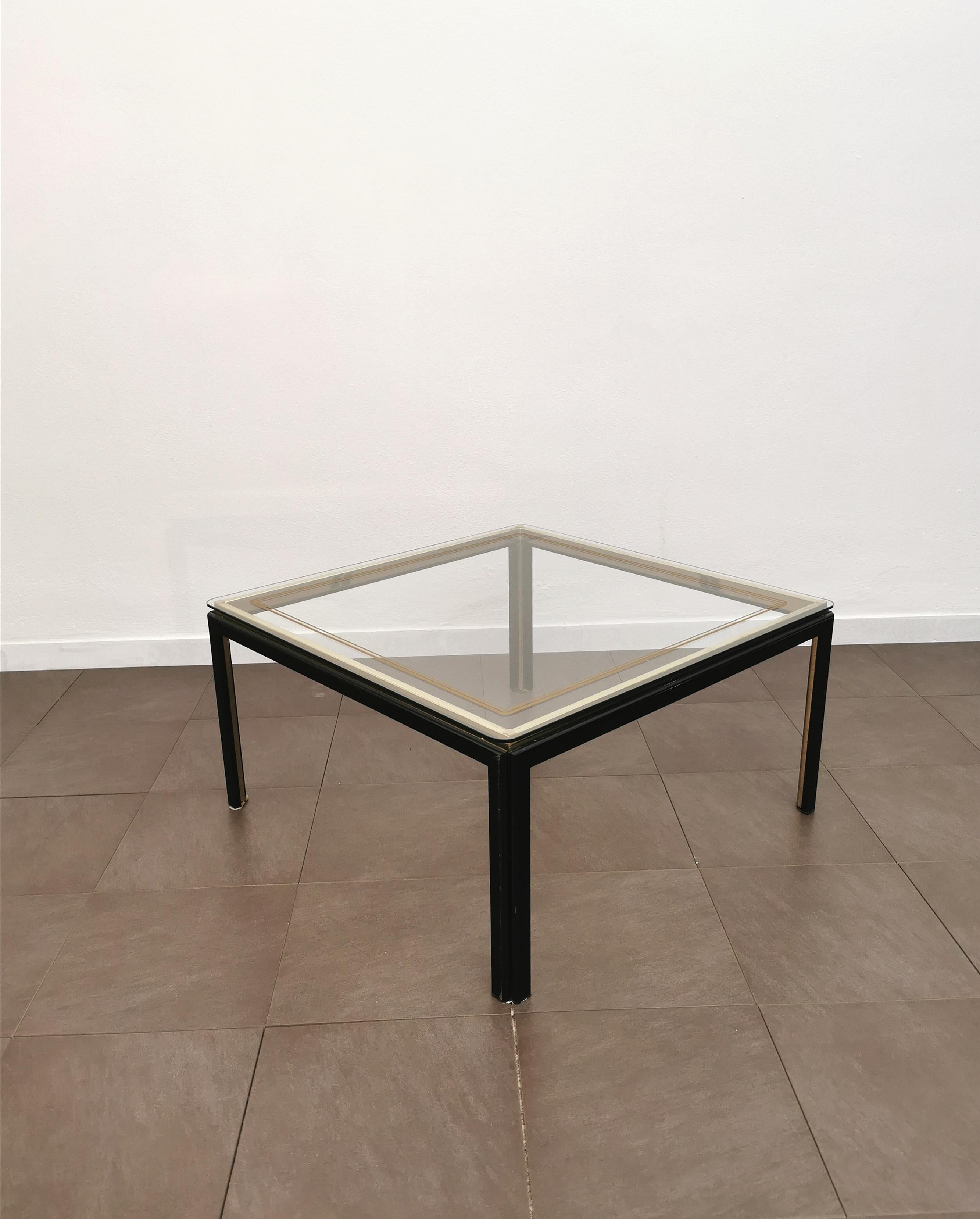 Coffee or sofa table produced in Italy in the 80s. The coffee table has a quadripod structure in black and golden enamelled metal and a square-shaped transparent glass top with rounded corners.