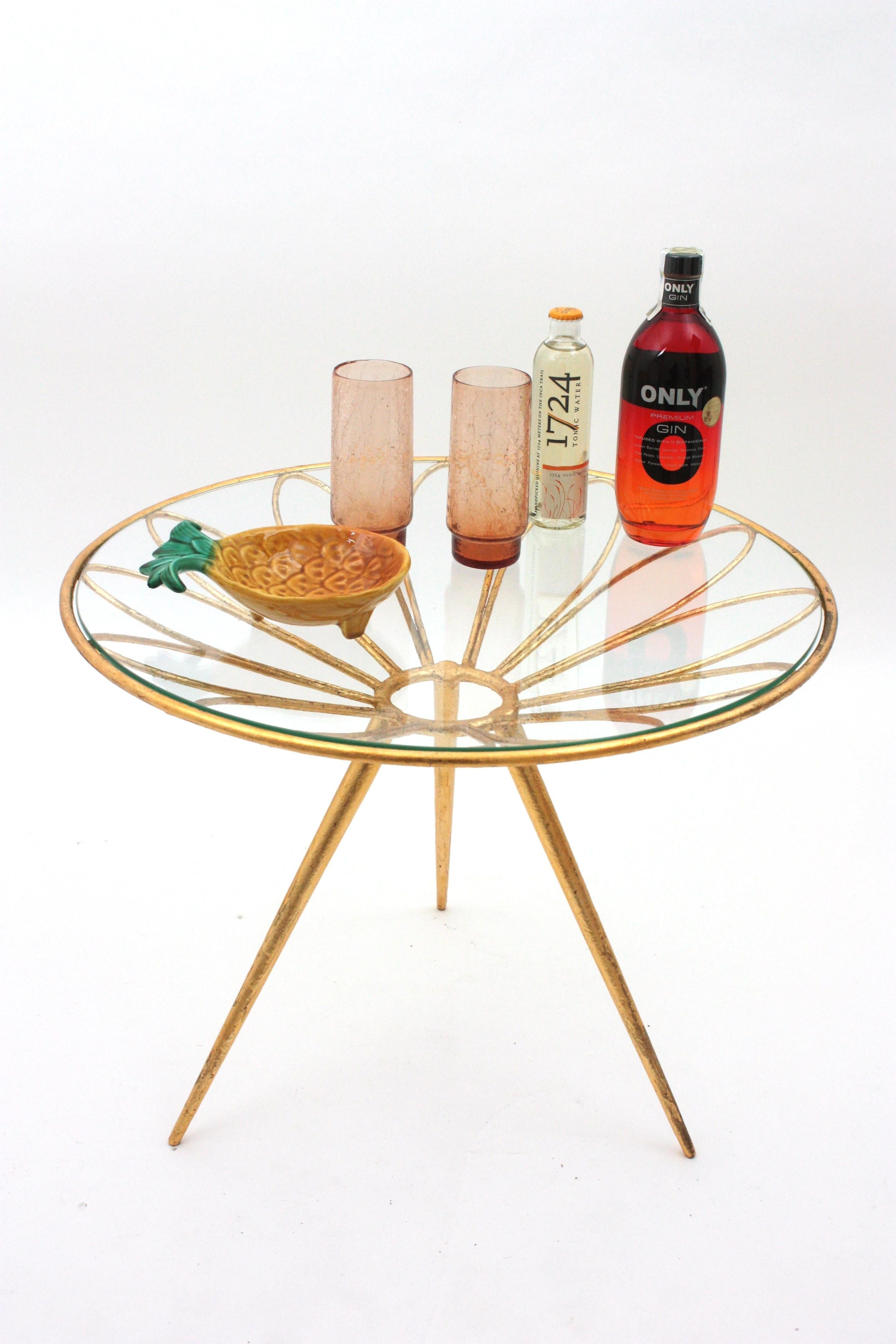 Table basse en fer doré à fleurs de marguerite, France, années 1950.
Table d'appoint glamour en fer doré à la feuille d'or avec plateau rond en verre reposant sur une base tripode. 
Cette table de cocktail présente une fleur de marguerite tenant un