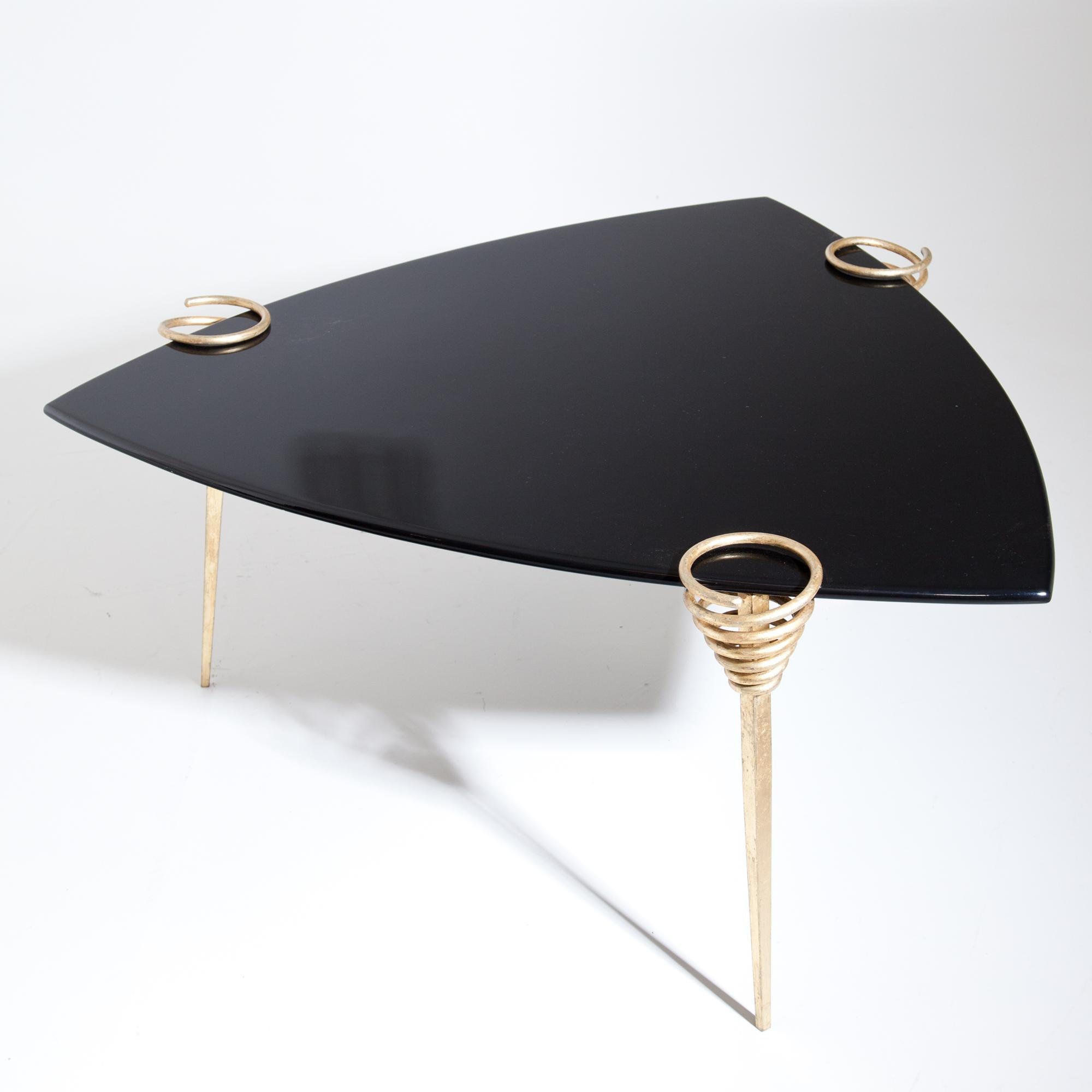 Coffee table with black triangular tabletop with convex sides standing on three legs. These are gold patinated and end in spirals that hold the tabletop.