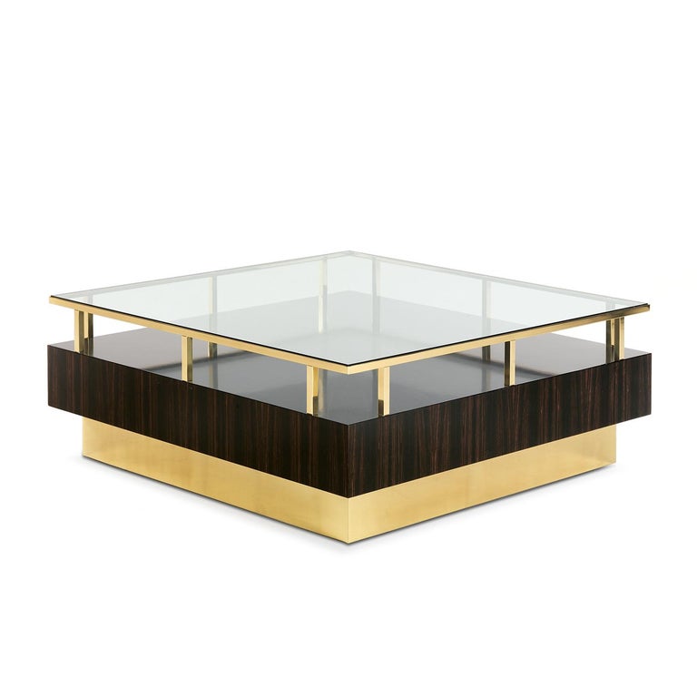 This elegant coffee table is part of a series of pieces with ebony as main element. The square base is in brass, and the luxurious hue of the metal strikingly complements the dark shade of the wood above. The table is adorned with a glass top
